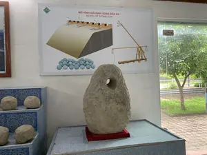 Bên trong một phòng trưng bày, có thể là một bảo tàng. Trên tường có treo một tấm áp phích với dòng chữ: "MÔ HÌNH GIẢ ĐỊNH SÚNG BẮN ĐÁ" và "MODEL OF STONE GUN". Tấm áp phích này minh họa mô hình của một loại máy bắn đá (súng thần công) được đặt trên tường thành và một bản vẽ chi tiết về cấu trúc của nó. Ở phía trước, một hiện vật bằng đá lớn, không đều, có một lỗ ở giữa, được trưng bày trên một bệ đỡ. Bên trái là các viên đạn đá hình cầu được đặt trên một kệ nhiều tầng có hoa văn màu xanh. Bên phải là một cửa kính nhìn ra không gian bên ngoài với cây cối và sân cỏ.。（日本語訳）。ある展示室内、おそらく博物館の中。壁には「MÔ HÌNH GIẢ ĐỊNH SÚNG BẮN ĐÁ」と「MODEL OF STONE GUN」という文字が書かれたポスターが掛けられている。このポスターは、城壁の上に設置された一種の投石機（神功砲）の模型と、その構造の詳細な図解を示している。手前には、中央に穴が開いた、大きくて不揃いな石の遺物が台座の上に展示されている。