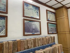 Đây là một phòng trưng bày, có thể là một phần của bảo tàng. Trên bức tường trắng có treo nhiều bức ảnh tư liệu được đóng trong khung gỗ. Các bức ảnh này ghi lại cảnh khai quật khảo cổ, cho thấy các hố đào, mặt cắt địa tầng và các nhà khảo cổ đang làm việc. Dưới các bức ảnh, trên một bệ trưng bày có hoa văn màu xanh và trắng, là những hàng gạch cổ, có thể là hiện vật được tìm thấy từ các cuộc khai quật. Một số tấm biển chú thích nhỏ màu xanh được đặt dưới các bức ảnh. Có thể đọc được nội dung của hai tấm biển: 'Khai quật di tích đường Hoàng Gia (2003)' và 'Mặt cắt hố khai quật La Thành (2008)'.。（日本語訳）。これは展示室で、博物館の一部かもしれません。白い壁には、木製の額縁に入れられた多くの資料写真が飾られています。写真の下、青と白の模様がある展示台の上には、古いレンガが並べられており、これらは発掘調査で発見された遺物である可能性があります。写真の下には、いくつかの小さな青いキャプションプレートが置かれています。2つのプレートの内容を読むことができます：「ホアンザー通り遺跡の発掘（2003年）」と「ラータイン発掘坑の断面（2008年）。