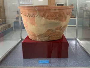 Trên bảng thông tin màu xanh lam có ghi: "Thống. Đất nung, triều Trần - Hồ, thế kỷ 14 - 15".。Bối cảnh là một bảo tàng hoặc phòng trưng bày. Trong ảnh là một chiếc thống lớn bằng đất nung, một hiện vật khảo cổ, được đặt trong tủ kính. Chiếc thống này có niên đại từ triều Trần - Hồ (thế kỷ 14-15), đã được phục dựng từ nhiều mảnh vỡ, với các đường nối được trám lại. Phần miệng thống có các họa tiết trang trí. Hiện vật được đặt trên một bệ đỡ hình hộp chữ nhật được bọc vải đỏ. Sàn nhà có màu xám xanh. Phía sau có thể thấy các hiện vật khác và phản chiếu của người xem trên mặt kính.。（日本語訳）。青い案内板には「壺。テラコッタ、陳-胡朝、14-15世紀」と書かれている。背景は博物館または展示室である。写真には、ガラスケースの中に置かれた、テラコッタ製の大きな壺、考古学的な遺物がある。この壺は陳-胡朝（14-15世紀）のもので、多くの破片から修復されており、接合部は埋められている。壺の口の部分には装飾的な模様がある。床は青みがかった灰色である。