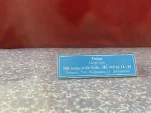 Hình ảnh chụp cận cảnh một tấm biển chú thích cho hiện vật, có thể ở trong một bảo tàng hoặc khu trưng bày. Tấm biển màu xanh lam với chữ trắng, được đặt trên một bề mặt màu xám có hoa văn. Phía sau là một vật thể lớn màu đỏ nâu. Nội dung trên tấm biển được ghi bằng tiếng Việt và tiếng Anh như sau:。"Thống。Large vase。Đất nung, triều Trần - Hồ, thế kỷ 14 - 15。Terracotta, Tran - Ho dynasties, 14 - 15th centuries"。Thông tin này mô tả hiện vật là một chiếc thống (bình lớn) làm bằng đất nung, có niên đại từ thời nhà Trần - Hồ, thế kỷ 14-15.。（日本語訳）。博物館または展示エリアにあると思われる、展示物のキャプションプレートのクローズアップ写真。青地に白文字のプレートは、模様のある灰色の表面に置かれています。背景には赤褐色の大きな物体があります。プレートの内容はベトナム語と英語で次のように書かれています：。「Thống（トン）。大きな壺。テラコッタ、陳・胡朝、14～15世紀。Terracotta, Tran - Ho dynasties, 14 - 15th centuries」。この情報は、展示物が14～15世紀の陳・胡朝時代に遡る、テラコッタで作られた大きな壺（thống）であることを説明しています。