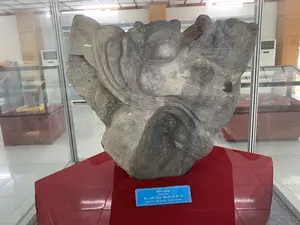 Bảng chú thích trên hiện vật ghi:。"Đầu rồng。Dragon Head。Đá, triều Trần - Hồ, thế kỷ 14 - 15。Stone, Tran - Ho dynasties, 14-15th centuries"。Bối cảnh là một không gian trưng bày trong nhà, có khả năng là một bảo tàng. Hiện vật chính là một tác phẩm điêu khắc bằng đá hình đầu rồng, có niên đại từ thế kỷ 14-15, thời Trần - Hồ. Đầu rồng được đặt trên một bệ đỡ được bọc vải đỏ và được bảo vệ bên trong một tủ trưng bày bằng kính. Phía sau có thể thấy các tủ trưng bày khác và một chiếc máy điều hòa không khí. Sàn nhà được lát gạch men màu trắng.。（日本語訳）。展示品の説明プレートには次のように書かれている：。「龍の頭。Dragon Head。石、陳・胡朝、14～15世紀。Stone, Tran - Ho dynasties, 14-15th centuries」。背景は屋内の展示スペースで、博物館である可能性が高い。主な展示品は、陳・胡朝時代の14～15世紀に遡る、龍の頭の形をした石の彫刻である。龍の頭は、赤い布で覆われた台座の上に置かれ、ガラスの展示ケースの中に保護されている。床は白いセラミックタイルで敷き詰められている。