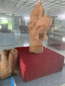 Bức ảnh chụp bên trong một không gian trưng bày, có khả năng là một bảo tàng. Đối tượng chính là một tác phẩm điêu khắc lớn bằng đất nung, mô tả đầu của một con chim thần thoại, có thể là đầu phượng hoàng, được đặt trong một tủ kính. Cổ vật này được đặt trên một bệ vuông phủ vải đỏ. Tác phẩm điêu khắc có các chi tiết tinh xảo như mào, mắt, và các lớp vảy trên cổ. Bên cạnh bệ trưng bày, dưới sàn nhà, là một mảnh vỡ khác của một cổ vật bằng đất nung. Có một bảng thông tin nhỏ màu xanh lam đặt gần mảnh vỡ, nhưng chữ trên đó không thể đọc rõ. Phía sau, có thể nhìn thấy các tủ trưng bày khác, các bức ảnh được đóng khung treo trên tường, và sàn nhà được lát gạch men sáng bóng. Không có người nào xuất hiện trong ảnh, và hoạt động chính là trưng bày các hiện vật lịch sử.。（日本語訳）。展示スペース、おそらく博物館の内部で撮影された写真です。主な被写体は、ガラスケースの中に置かれた、神話上の鳥、おそらく鳳凰の頭をかたどった大きなテラコッタの彫刻です。この遺物は、赤い布で覆われた四角い台座の上に置かれています。彫刻には、とさか、目、首の鱗の層など、精巧なディテールがあります。展示台の横の床には、別のテラコッタの遺物の破片があります。破片の近くに小さな青い案内板が置かれていますが、その上の文字ははっきりと読み取れません。後方には、他の展示ケース、壁に掛けられた額入りの写真、そして光沢のあるタイル張りの床が見えます。写真には誰も写っておらず、主な活動は歴史的遺物の展示です。