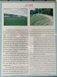 Đây là hình ảnh một tấm bảng thông tin giới thiệu về di tích La Thành, một phần của Di sản Thế giới Thành Nhà Hồ. Tấm bảng có nền trắng, khung viền hoa văn màu xanh lam, trình bày nội dung song ngữ Việt - Anh.。Nội dung văn bản tiếng Việt trên bảng được ghi lại như sau:。LA THÀNH。La thành được xây dựng tháng 9 năm 1399, là vòng thành ngoài bảo vệ toàn bộ các kiến trúc và cư dân trong kinh thành. La thành dài khoảng 10km, nằm cách hoàng thành khoảng 1-3km. Nhiều đoạn được xây dựng đào đắp bằng đất dựa theo địa hình tự nhiên, kết nối với các quả núi quanh thành, chạy từ núi Đốn Sơn (xã Vĩnh Thành) đến núi Hắc Khuyển (xã Vĩnh Long), các núi Xuân Đài, Trác Phong, Tiến Sỹ (xã Vĩnh Ninh), núi An Tôn (xã Vĩnh Yên), Thổ Tượng (xã Vĩnh Quang). Trong đó có nhiều đoạn ven theo sông Bưởi và sông Mã.。Đoạn La thành hiện còn khá nguyên vẹn tại xã Vĩnh Phúc (phía Đông Nam Thành Nhà Hồ) và cánh đồng xã Vĩnh Long (phía Đông Bắc Thành Nhà Hồ). Kết quả thám sát khảo cổ học ở đây cho thấy mặt cắt La thành hình thang, cao 6m, chân thành rộng 37m, mặt thành rộng khoảng 9,20m.。Di tích được Bộ Văn hóa, Thể thao và Du lịch xếp hạng Di tích Lịch sử Văn hóa Quốc gia năm 2011 và là bộ phận quan trọng trong cấu trúc tổng thể của Di sản Thế giới Thành Nhà Hồ.。Ngoài văn bản, trên bảng còn có hai bức ảnh minh họa:。1. Ảnh bên trái: Một cánh đồng lúa xanh rộng lớn, phía xa là hàng cây dưới bầu trời có mây.。2. Ảnh bên phải: Một đoạn của La Thành, thể hiện một bờ thành bằng đất phủ cỏ xanh với một con đường mòn trên đỉnh, xung quanh là cây cối rậm rạp.。Không có hoạt động nào của con người được ghi nhận, đây là một bảng thông tin tĩnh.。（日本語訳）。これは、世界遺産である胡朝の城塞の一部である羅城遺跡を紹介する案内板の写真です。案内板は白い背景に青い模様の縁取りがあり、ベトナム語と英語の二か国語で内容が記載されています。案内板に記載されているベトナム語の文章は以下の通りです。羅城。羅城は1399年9月に建設され、都城内のすべての建築物と住民を保護する外側の城壁です。羅城の長さは約10kmで、皇城から約1～3kmの場所に位置しています。多くの区間は自然の地形に基づいて土を掘り固めて建設され、城の周りの山々を結び、ドンソン山（ヴィンタイン社）からハックエン山（ヴィンロン社）、スアンダイ山、チャックフォン山、ティエンシー山（ヴィンニン社）、アントン山（ヴィンイエン社）、トートゥオン山（ヴィンクアン社）まで続いています。その中には、ブオイ川とマー川に沿った多くの区間が含まれています。羅城の区間は現在、ヴィンフック社（胡朝の城塞の南東）とヴィンロン社の田園地帯（胡朝の城塞の北東）でかなり完全な状態で残っています。ここでの考古学調査の結果、羅城の断面は台形で、高さ6m、城壁の基部の幅は37m、城壁上部の幅は約9.20mであることが示されています。この遺跡は2011年に文化・スポーツ・観光省によって国家歴史文化遺跡に指定され、世界遺産である胡朝の城塞の全体構造における重要な部分です。文章の他に、案内板には2枚のイラスト写真があります。1. 左の写真：広大な緑の田んぼ、遠くには曇り空の下に並木が見えます。2. 右の写真：羅城の一区間で、緑の草で覆われた土の城壁とその頂上にある小道、周りは鬱蒼とした木々が描かれています。人間の活動は記録されておらず、これは静的な案内板です。
