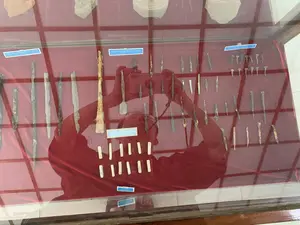 Bức ảnh chụp một tủ trưng bày hiện vật trong một bảo tàng hoặc khu di tích. Bên trong tủ kính có khung gỗ, trên nền vải màu đỏ, là các hiện vật khảo cổ. Các hiện vật bao gồm nhiều mũi tên sắt, đinh sắt với các kích cỡ khác nhau, và các miếng chì lưới hình chữ nhật. Các đồ vật bằng kim loại này có dấu hiệu bị oxy hóa và gỉ sét. Có các nhãn nhỏ màu xanh ghi chú thích bằng tiếng Việt và tiếng Anh cho các nhóm hiện vật.。Nội dung văn bản trên các nhãn ghi chú:。- "Mũi tên sắt, khai quật Thành Nhà Hồ và đàn tế Nam Giao / Iron arrows, excavated at the Ho Citadel site and Nam Giao altar"。- "Đinh sắt, khai quật Thành Nhà Hồ và đàn tế Nam Giao / Iron nails, excavated at the Ho Citadel site and Nam Giao altar"。- "Chì lưới / Lead fishing nets"。Hoạt động chính là trưng bày các hiện vật lịch sử. Trên mặt kính của tủ có phản chiếu rõ hình ảnh một người đang dùng điện thoại thông minh để chụp lại các hiện vật.。（日本語訳）。写真は、博物館または史跡にある遺物の展示ケースを写したものです。木製のフレームが付いたガラスケースの中、赤い布地の上に、考古学的な遺物があります。遺物には、多くの鉄の矢じり、さまざまなサイズの鉄釘、そして長方形の鉛の網おもりが含まれています。これらの金属製の品々には、酸化や錆の痕跡が見られます。遺物のグループごとに、ベトナム語と英語で注釈が書かれた小さな青いラベルがあります。注釈ラベルのテキスト内容：。- 「鉄の矢じり、胡朝の城塞及び南郊壇にて発掘 / Iron arrows, excavated at the Ho Citadel site and Nam Giao altar」。- 「鉄釘、胡朝の城塞及び南郊壇にて発掘 / Iron nails, excavated at the Ho Citadel site and Nam Giao altar」。- 「網おもり / Lead fishing nets」。主な活動は、歴史的な遺物を展示することです。ケースのガラス面には、スマートフォンを使って遺物を撮影している人物の姿がはっきりと映り込んでいます。