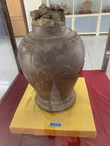 Bức ảnh chụp một hiện vật khảo cổ được trưng bày trong tủ kính, có thể là ở một bảo tàng. Hiện vật chính là một chiếc hũ sành cổ, lớn, màu nâu sẫm. Trên thân hũ có các hoa văn nổi, bao gồm họa tiết hình mây và các hoa văn trang trí ở phần vai và đế hũ. Bên trong miệng hũ chứa đầy các vật thể hình trụ, trông giống như những cuộn tiền xu cổ đã bị oxy hóa và kết dính lại với nhau. Chiếc hũ được đặt trên một bệ trưng bày được bọc vải màu vàng, trên một bề mặt phủ vải đỏ. Phía trước hũ có một tấm biển nhỏ màu xanh ghi chữ trắng: "Hũ tiền đồng" và dòng chữ tiếng Anh "Copper coins". Phía sau là một tủ trưng bày khác có cửa kính, bên trong có thể thấy các hiện vật gốm khác.。（日本語訳）。この写真は、博物館と思われる場所のガラスケースに展示されている考古学的遺物を撮影したものです。主な遺物は、大きくて濃い茶色の古い陶器の壺です。壺の胴体には、雲の模様や、壺の肩と底の部分の装飾模様を含む、浮き彫りの文様があります。その壺は、赤い布で覆われた台座の上にある、黄色い布で覆われた展示台の上に置かれています。壺の前には、「Hũ tiền đồng」と英語の「Copper coins」という白い文字が書かれた小さな青いプレートがあります。後ろにはガラス扉のついた別の展示ケースがあり、その中には他の陶器の遺物が見えます。