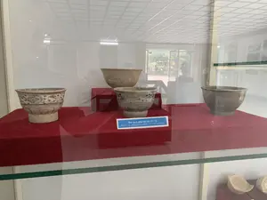 Bối cảnh là một phòng trưng bày hoặc bảo tàng. Bên trong một tủ kính, có bốn chiếc bát gốm cổ đang được trưng bày trên các bệ đỡ bọc vải đỏ. Một số bát có màu trơn, trong khi những chiếc khác được trang trí hoa văn màu xanh lam trên nền men trắng. Phía trước có một tấm biển nhỏ ghi chú thông tin về hiện vật. Văn bản trên biển ghi: "Bát men, gốm thế kỷ 15 - 16" và "Bowl with white glaze, ceramic 15th - 16th centuries". Ở kệ dưới của tủ kính, có thể thấy một vài mảnh gốm vỡ. Phía sau, qua lớp kính, có sự phản chiếu của trần nhà và không gian bên ngoài với cây cối.。（日本語訳）。背景は展示室または博物館です。ガラスケースの中には、赤い布で覆われた台座の上に4つの古い陶磁器の碗が展示されています。いくつかの碗は無地ですが、他のものは白い釉薬の地に青い模様で装飾されています。手前には、展示物に関する情報を記した小さなプレートがあります。プレートには「Bát men, gốm thế kỷ 15 - 16」および「Bowl with white glaze, ceramic 15th - 16th centuries」と記されています。ガラスケースの下の棚には、いくつかの割れた陶器の破片が見えます。後ろでは、ガラスに天井や木々のある屋外の空間が映り込んでいます。
