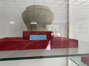 Bức ảnh chụp một hiện vật khảo cổ đang được trưng bày trong một tủ kính, có khả năng là tại một bảo tàng hoặc phòng triển lãm. Hiện vật là một chiếc vò gốm cổ, màu xanh xám, có nhiều vết nứt, được đặt trên một bệ đỡ bọc vải màu đỏ. Phía trước hiện vật có một tấm biển nhỏ màu xanh lam ghi chú thích bằng tiếng Việt và tiếng Anh. Dòng chữ tiếng Việt là 'ĐỒ ĐỰNG, THẾ KỶ 14 - 15'. Dòng chữ tiếng Anh là 'Jars, 14th - 15th centuries'. Bề mặt kính của tủ trưng bày phản chiếu hình ảnh trần nhà dạng ô lưới, đèn chiếu sáng và không gian xung quanh phòng trưng bày.。（日本語訳）。写真は、ガラスケースの中に展示されている考古学的遺物を写したもので、おそらく博物館か展示室のものです。遺物は、灰青色で多くのひび割れがある古い陶器の壺で、赤い布で覆われた台座の上に置かれています。遺物の前には、ベトナム語と英語で説明が書かれた青色の小さなプレートがあります。ベトナム語の表記は「ĐỒ ĐỰNG, THẾ KỶ 14 - 15」です。英語の表記は「Jars, 14th - 15th centuries」です。展示ケースのガラス面には、格子状の天井、照明、そして展示室の周囲の空間が反射しています。