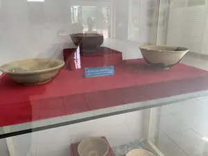 Bên trong một bảo tàng hoặc phòng trưng bày, có một tủ kính trưng bày các hiện vật. Bên trong tủ, trên một bề mặt được phủ vải đỏ, có ba chiếc bát gốm cổ được trưng bày. Một chiếc bát màu nâu sẫm được đặt trên một bệ trưng bày màu đỏ ở phía sau. Hai chiếc bát khác, một màu be và một màu be với các mảng tối, được đặt ở phía trước. Giữa các hiện vật có một tấm biển nhỏ màu xanh ghi chú thích bằng tiếng Việt và tiếng Anh. Nội dung trên biển là: "SƯU TẬP BÁT, THẾ KỶ 14 - 15" và "Collection of various types of bowls, 14th - 15th centuries". Phía sau tủ kính là không gian của bảo tàng với sàn gạch trắng và cửa kính. Có hình ảnh phản chiếu trên mặt kính của tủ trưng bày.。（日本語訳）。博物館または展示室の中に、展示品を陳列するガラスケースがあります。ケースの中、赤い布で覆われた表面の上に、3つの古い陶器の碗が展示されています。濃い茶色の碗が一つ、後方の赤い展示台の上に置かれています。他の2つの碗、一つはベージュ色、もう一つは暗い斑点のあるベージュ色のものが、前方に置かれています。展示品の間には、ベトナム語と英語で説明が書かれた小さな青いプレートがあります。プレートの内容は、「SƯU TẬP BÁT, THẾ KỶ 14 - 15」および「Collection of various types of bowls, 14th - 15th centuries」です。ガラスケースの後ろは、白いタイルの床とガラスのドアがある博物館の空間です。展示ケースのガラス面には、反射した像があります。
