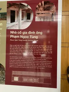 Bức ảnh chụp một bảng thông tin màu đỏ sẫm giới thiệu về một công trình kiến trúc. Trên bảng có tiêu đề lớn bằng chữ trắng: "Nhà cổ gia đình ông Phạm Ngọc Tùng", và dòng chữ tiếng Anh "Pham Ngoc Tung Family Ancient House".。Nội dung chi tiết bằng tiếng Việt trên bảng như sau: "Đây là ngôi nhà cổ dân gian được các chuyên gia đánh giá cao bởi kiến trúc đặc sắc. Ngôi nhà được xây dựng vào năm 1810, dưới bàn tay của những người thợ tài hoa. Ngôi nhà có bảy gian bằng gỗ, kiến trúc theo kiểu chồng rường, kẻ chuyền và chồng rường kẻ bẩy. Hoa văn trang trí được chạm trổ rất tinh xảo, thể hiện sinh động các chủ đề tứ linh (long, ly, quy, phượng), tứ quý (tùng, cúc, trúc, mai).".。Bảng thông tin này có hình ảnh minh họa là các bức ảnh chụp chi tiết kiến trúc bên trong ngôi nhà gỗ cổ và một bản vẽ kiến trúc của ngôi nhà. Bối cảnh cho thấy bảng thông tin được đặt trong một không gian trong nhà, có thể là bảo tàng hoặc khu trưng bày, với bức tường ốp gỗ phía sau và một phần của chiếc quạt điện màu đen ở bên phải.。（日本語訳）。この写真は、ある建築物を紹介する濃い赤色の案内板を写したものです。案内板には白い大きな文字で「ファム・ゴック・トゥン氏の旧家」という見出しと、英語で「Pham Ngoc Tung Family Ancient House」という表記があります。案内板に記載されているベトナム語の詳細は以下の通りです。「これは、その独特な建築様式により専門家から高く評価されている民間の古民家です。この家は1810年に、才能ある職人たちの手によって建てられました。この家は木造七間で、チョン・ルオン、ケ・チュエン、チョン・ルオン・ケ・バイ様式で建てられています。装飾文様は非常に精巧に彫られており、四霊（龍、麒麟、亀、鳳凰）、四君子（松、菊、竹、梅）といったテーマを生き生きと表現しています。」。この案内板には、古い木造家屋の内部の建築詳細を写した写真や、その家の建築図面が説明画像として添えられています。背景から、この案内板が屋内の空間、おそらく博物館か展示エリアに設置されており、後ろには板張りの壁、右側には黒い扇風機の一部が見えていることがわかります。