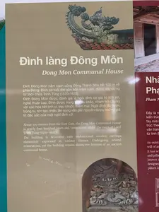 Đây là hình ảnh chụp một bảng thông tin giới thiệu về di tích lịch sử. Bảng thông tin này cung cấp chi tiết về 'Đình làng Đông Môn' bằng cả tiếng Việt và tiếng Anh.。Nội dung chữ viết trên bảng:。Tiêu đề: "Đình làng Đông Môn", "Dong Mon Communal House".。Nội dung tiếng Việt: "Đình Đông Môn nằm cách cổng Đông Thành Nhà Hồ 100 m về phía Đông. Đình có tuổi đời gần bốn trăm năm, được xây dựng từ thời chúa Trịnh Tùng (1570-1623). Đình Đông Môn được đánh giá là ngôi đình có giá trị lịch sử, nghệ thuật cao. Đình được trang trí điêu khắc, chạm trổ cầu kỳ với các họa tiết tinh vi, trau chuốt, mềm mại. Ngôi đình đã được trùng tu, tôn tạo nhiều lần song vẫn giữ nguyên được những giá trị đặc sắc của một ngôi đình cổ."。Cảnh vật và đồ vật:。Bảng thông tin được đặt tại một khu di tích, có thể là trong khuôn viên Thành Nhà Hồ. Trên bảng có các hình ảnh minh họa cho ngôi đình, bao gồm:。- Một bức ảnh chụp chi tiết phần đầu đao trên mái ngói, được chạm khắc công phu hình đầu rồng.。- Một bức ảnh chụp cận cảnh các tác phẩm chạm khắc gỗ phức tạp bên trong kiến trúc đình.。- Một bản vẽ phác thảo kiến trúc tổng thể của ngôi đình.。Bên phải, có một phần của một bảng thông tin khác màu đỏ sẫm, giới thiệu về một địa điểm khác bắt đầu bằng "Nhà Pha...", nhưng phần lớn nội dung đã bị che khuất và không thể đọc được.。（日本語訳）。これは歴史的遺跡を紹介する案内板を撮影した画像です。この案内板は、「ドンモン村の亭」に関する詳細をベトナム語と英語の両方で提供しています。案内板に書かれている内容：。タイトル：「Đình làng Đông Môn」、「Dong Mon Communal House」。ベトナム語の内容：「ドンモン亭は胡朝の城の東門から東へ100mの場所にあります。この亭は約400年の歴史があり、鄭松（チン・トゥン）領主（1570-1623）の時代に建てられました。ドンモン亭は、歴史的、芸術的に高い価値を持つ亭であると評価されています。亭は、精巧で洗練された、柔らかいモチーフで、凝った彫刻や彫刻で装飾されています。この亭は何度も修復・改修されましたが、古い亭の独特な価値を今もなお保っています。」。風景と物：。案内板は遺跡内に設置されており、胡朝の城の敷地内である可能性があります。案内板には、亭を説明する以下の画像が含まれています：。- 瓦屋根の棟の端の部分（đầu đao）を詳細に撮影した写真。龍の頭の形に精巧に彫刻されています。- 亭の建築内部にある複雑な木彫り作品のクローズアップ写真。- 亭の全体的な建築のスケッチ図。右側には、濃い赤色の別の案内板の一部があり、「Nhà Pha...」で始まる別の場所を紹介していますが、内容の大部分は隠れていて読むことができません。