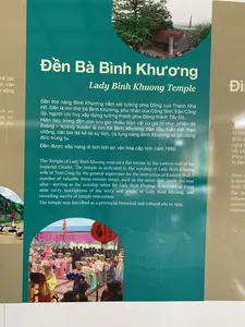 Đây là hình ảnh một tấm biển thông tin giới thiệu về di tích lịch sử Đền Bà Bình Khương tại khu vực Thành Nhà Hồ.。**1. Thông tin văn bản được ghi lại:**。- Tiêu đề: Đền Bà Bình Khương (Lady Binh Khuong Temple).。- Nội dung chính: "Đền thờ nàng Bình Khương nằm sát tường phía Đông của Thành Nhà Hồ. Đền là nơi thờ bà Bình Khương, phu nhân của Cống Sinh Trần Công Sỹ, người chỉ huy xây dựng tường thành phía Đông thành Tây Đô. Hiện nay, trong đền còn lưu giữ nhiều hiện vật có giá trị như: phiến đá thiêng – tương truyền là nơi bà Bình Khương đập đầu tuẫn tiết theo chồng, các bia đá kể lại sự tích, ca tụng nàng Bình Khương và ghi công đức trùng tu. Đền được xếp hạng di tích lịch sử văn hóa cấp tỉnh năm 1995."。**2. Mô tả cảnh vật và hoạt động:**。- **Địa điểm:** Tấm biển thông tin này giới thiệu về một ngôi đền. Dựa trên văn bản, đây là Đền Bà Bình Khương, nằm gần Thành Nhà Hồ.。- **Hoạt động và vật thể:** Tấm biển trưng bày nhiều hình ảnh khác nhau.。- Một bức ảnh chụp kiến trúc bên ngoài của ngôi đền, là một công trình cổ có mái ngói, nằm trong một khoảng sân có cây cối.。- Một bức ảnh khác ghi lại cảnh một lễ hội hoặc nghi lễ văn hóa. Trong ảnh, nhiều người, chủ yếu là phụ nữ, mặc trang phục truyền thống (áo dài, áo tứ thân) nhiều màu sắc. Một người mặc áo dài màu vàng dường như đang chủ trì buổi lễ. Phía trên có treo một chiếc đèn lồng đỏ. Đông đảo người dân đang đứng xem ở phía sau.。- Một bức ảnh nhỏ hơn chụp một người đứng gần một bức tường thành bằng đá lớn và một đàn ngỗng.。- Ngoài ra còn có một bản vẽ phác thảo kiến trúc của ngôi đền.。（日本語訳）。これは、胡朝の城塞地域にある歴史的遺跡、ビン・クオン夫人祠に関する案内板の写真です。**1. 記録されている文章の情報：**。- タイトル：ビン・クオン夫人祠（Lady Binh Khuong Temple）。- 主な内容：「ビン・クオン夫人を祀る祠は、胡朝の城塞の東側の壁に隣接しています。この祠は、西都城の東側の城壁建設を指揮した貢生チャン・コン・シーの夫人であるビン・クオン夫人を祀る場所です。現在、祠の中には多くの貴重な遺物が保存されています。例えば、聖なる石板（ビン・クオン夫人が夫に殉じて頭を打ち付けたと伝えられる場所）、事績を語り、ビン・クオン夫人を称え、修復の功徳を記録した石碑などです。この祠は1995年に省レベルの歴史文化遺跡に指定されました。」。**2. 風景と活動の説明：**。- **場所：** この案内板は一つの祠について紹介しています。文章に基づくと、これは胡朝の城塞の近くにあるビン・クオン夫人祠です。- **活動と物体：** 案内板には様々な写真が展示されています。- 祠の外観を撮影した写真が1枚。これは瓦屋根を持つ古い建築物で、木々のある庭の中にあります。- 祭りまたは文化的な儀式の様子を記録した別の写真が1枚。写真には、多くの人々、主に女性が、色とりどりの伝統衣装（アオザイ、アオトゥーザン）を着用しています。黄色いアオザイを着た一人が儀式を主宰しているようです。上には赤い提灯が吊るされています。多くの人々が後ろに立って見ています。- 大きな石の城壁とガチョウの群れの近くに立つ人を撮影した、より小さな写真が1枚。- その他に、祠の建築スケッチ図もあります。