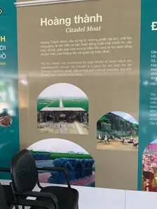 Đây là hình ảnh một tấm bảng thông tin được trưng bày trong nhà, có khả năng là tại khu di tích hoặc bảo tàng Thành nhà Hồ. Phía trước có một chiếc ghế màu đen. Tấm bảng cung cấp thông tin về 'Hoàng thành'.。Nội dung văn bản có thể đọc được trên bảng:。- Tiêu đề: 'Hoàng thành', 'Citadel Moat'.。- Đoạn mô tả bằng tiếng Việt: 'Hoàng Thành được xây dựng từ những phiến đá lớn, chế tác công phu, là nơi diễn ra các hoạt động thiết chế chính trị, văn hóa, xã hội, giáo dục của vương triều Hồ, xưa là nơi sinh sống và làm việc của hoàng tộc và quan lại triều đình.'。- Các chữ có thể nhìn thấy ở các phần khác của bảng: 'H', 'ỚI', 'HỒ', 'Site' (bên trái) và 'Đền', 'Hồ.', 'Sỹ,' (bên phải).。Các hình ảnh trên bảng mô tả:。- Một cổng thành cổ bằng đá với các lối đi hình vòm, phía sau là con đường thẳng tắp chạy giữa cánh đồng lúa xanh.。- Một đàn ngỗng đang đi trên đường đất bên cạnh một bức tường thành bằng đá.。- Một tác phẩm điêu khắc đá hình rồng trên bệ gạch, phía sau là cánh đồng.。- Một phần hình ảnh cho thấy những người đang tham gia một nghi lễ trong trang phục truyền thống màu vàng và đỏ.。（日本語訳）。これは屋内に展示されている案内板の写真で、おそらく胡朝の城の遺跡または博物館にあるものです。手前には黒い椅子が1脚あります。案内板は「皇城」に関する情報を提供しています。案内板で読み取れる文章の内容：。- 見出し：「皇城」、「Citadel Moat」。- ベトナム語による説明文：「皇城は、精巧に加工された大きな石板から建てられ、胡朝の政治、文化、社会、教育の制度的活動が行われた場所であり、かつては皇族と朝廷の役人が生活し、働く場所でした。」。案内板に描かれている写真：。- アーチ型の通路がある石造りの古い城門、その後ろには青々とした田んぼの間をまっすぐに走る道。- 石垣の横の土道を歩いているガチョウの群れ。- 煉瓦の台座の上にある龍の石の彫刻、その後ろは野原。- 黄色と赤の伝統的な衣装を着て儀式に参加している人々を示す写真の一部。