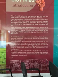 Đây là hình ảnh chụp một tấm bảng thông tin giới thiệu về Thành Nhà Hồ, có thể được đặt trong một bảo tàng hoặc khu trưng bày tại di tích. Phía trước tấm bảng có hai chiếc ghế văn phòng màu đen. Tấm bảng có nền màu đỏ sẫm, chứa đựng thông tin bằng cả tiếng Việt và tiếng Anh.。Nội dung chữ viết tiếng Việt được ghi lại như sau:。"GIỚI THIỆU。Thành Nhà Hồ là kinh đô của nước Đại Việt thời nhà Trần (1398-1400) và nước Đại Ngu thời Nhà Hồ (1400-1407).。Thành được Hồ Quý Ly cho xây dựng năm Đinh Sửu (1397). Điểm độc đáo là tường thành và các cổng vòm cuốn được xây bằng những khối đá lớn, nặng từ 10 đến 26 tấn, được chế tác công phu, chồng xếp lên nhau chắc chắn mà không có chất kết dính nào.。Giá trị nổi bật của Thành Nhà Hồ không chỉ chứa đựng ở các di sản văn hóa vật thể như thành quách, công trình kiến trúc, vết tích hoàng cung, v.v. mà còn ở các giá trị di sản phi vật thể tạo nên chúng. Ngày 27/6/2011, UNESCO đã ghi danh Thành Nhà Hồ là Di sản Thế giới."。Ngoài ra, trên bảng còn có hình ảnh một hiện vật gốm trang trí hình đầu chim, một bản vẽ kỹ thuật mô tả cấu trúc cổng thành bằng đá, và một bức ảnh nhỏ về cánh đồng lúa.。（日本語訳）。案内板の前には黒いオフィスチェアが2脚あります。案内板は濃い赤色の背景で、ベトナム語と英語の両方で情報が記載されています。ベトナム語で書かれた内容は以下の通りです。「紹介。胡朝の城塞は、陳朝時代（1398-1400）の大越国、および胡朝時代（1400-1407）の大虞国の首都でした。城は丁丑（ていちゅう）の年（1397年）に胡季犛（ホー・クイ・リ）によって建設されました。そのユニークな点は、城壁とアーチ型の門が、重さ10トンから26トンの大きな石塊で造られていることです。それらは精巧に加工され、接着剤を一切使わずに頑丈に積み重ねられています。胡朝の城塞の際立った価値は、城郭、建築物、王宮の痕跡などの有形文化遺産だけでなく、それらを生み出した無形文化遺産の価値にも含まれています。2011年6月27日、ユネスコは胡朝の城塞を世界遺産に登録しました。」。その他、板上には鳥の頭の形をした装飾陶器の遺物の写真、石造りの城門の構造を示す技術図面、そして水田の小さな写真もあります。