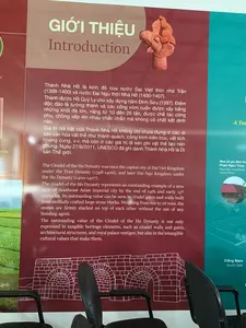 Bức ảnh chụp một tấm bảng thông tin lớn trong nhà, có thể là tại một bảo tàng hoặc trung tâm trưng bày di tích Thành Nhà Hồ. Tấm bảng có nền màu đỏ đô và xanh lam, chứa thông tin giới thiệu về di tích bằng cả tiếng Việt và tiếng Anh. Phía trước tấm bảng là lưng của hai chiếc ghế văn phòng màu đen.。Nội dung văn bản trên bảng như sau:。Tiêu đề chính:。"GIỚI THIỆU。Introduction"。Nội dung tiếng Việt:。"Thành Nhà Hồ là kinh đô của nước Đại Việt thời nhà Trần (1398-1400) và nước Đại Ngu thời Nhà Hồ (1400-1407). Thành được Hồ Quý Ly cho xây dựng năm Đinh Sửu (1397). Điểm độc đáo là tường thành và các cổng vòm cuốn được xây bằng những khối đá lớn, nặng từ 10 đến 26 tấn, được chế tác công phu, chồng xếp lên nhau chắc chắn mà không có chất kết dính nào. Giá trị nổi bật của Thành Nhà Hồ không chỉ chứa đựng ở các di sản văn hóa vật thể như thành quách, công trình kiến trúc, vết tích hoàng cung, v.v. mà còn ở các giá trị di sản phi vật thể tạo nên chúng. Ngày 27/6/2011, UNESCO đã ghi danh Thành Nhà Hồ là Di sản Thế giới."。Nội dung tiếng Anh:。"The Citadel of the Ho Dynasty was once the capital city of Dai Viet Kingdom under the Tran Dynasty (1398-1400), and later Dai Ngu Kingdom under the Ho Dynasty (1400-1407). The citadel of the Ho Dynasty represents an outstanding example of a new style of Southeast Asian imperial city by the end of 14th and early 15th centuries. Its outstanding value can be seen in citadel gates and walls built from skillfully crafted large stone blocks. Weighing from ten to 26 tons, the stones are firmly stacked on top of each other without the use of any bonding agent. The outstanding value of the Citadel of the Ho Dynasty is not only expressed in tangible heritage elements, such as citadel walls and gates, architectural structures, and royal palace vestiges, but also in the intangible cultural values that make them."。Các chú thích khác trên bảng:。- "Nhà cổ gia đình ông Phạm Ngọc Tùng / Pham Ngoc Tung Family Ancient House"。- "Cổng Nam / South Gate"。Ngoài văn bản, trên bảng còn có hình ảnh một hiện vật điêu khắc đầu chim bằng đất nung, một bản vẽ kỹ thuật của cổng thành, và các hình ảnh minh họa khác.。（日本語訳）。写真は屋内の大きな案内板を写したもので、おそらく胡朝の城跡の博物館または展示センターで撮影されたものです。案内板はワインレッドと青を背景とし、ベトナム語と英語の両方で遺跡に関する紹介情報が記載されています。案内板の前には、黒いオフィスチェア2脚の背もたれが見えます。案内板の文章内容は以下の通りです。メインタイトル：。「GIỚI THIỆU。Introduction」（紹介）。ベトナム語の内容：。「胡朝の城は、陳朝時代（1398-1400）の大越国、および胡朝時代（1400-1407）の大虞国の首都でした。城は丁丑の年（1397年）に胡季犛によって建設が命じられました。そのユニークな点は、城壁とアーチ型の門が、重さ10トンから26トンの大きな石塊で造られていることです。これらの石は精巧に加工され、接着剤を一切使わずに頑丈に積み重ねられています。胡朝の城の際立った価値は、城郭、建築物、王宮の痕跡などの有形文化遺産だけでなく、それらを創り出した無形文化遺産の価値にも含まれています。2011年6月27日、ユネスコは胡朝の城を世界遺産に登録しました。」。英語の内容：。「胡朝の城は、かつて陳朝（1398-1400）下の大越国の首都であり、その後、胡朝（1400-1407）下の大虞国の首都でした。胡朝の城は、14世紀末から15世紀初頭にかけての東南アジアの新しい様式の皇都の優れた例を表しています。その際立った価値は、巧みに作られた大きな石のブロックから建てられた城門と城壁に見ることができます。重さ10トンから26トンの石は、接着剤を一切使用せずに互いの上にしっかりと積み重ねられています。胡朝の城の際立った価値は、城壁や城門、建築構造物、王宮の痕跡などの有形遺産要素だけでなく、それらを生み出した無形文化価値にも表れています。」。案内板上のその他の注釈：。- 「ファム・ゴック・トゥン氏の古民家 / Pham Ngoc Tung Family Ancient House」。- 「南門 / South Gate」。文章の他に、案内板にはテラコッタ製の鳥の頭の彫刻の遺物、城門の技術図面、その他のイラスト画像もあります。