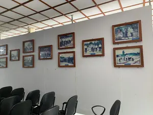 Bên trong một căn phòng, có thể là phòng trưng bày hoặc phòng họp. Trên bức tường trắng được treo rất nhiều bức ảnh tư liệu đã cũ, lồng trong khung gỗ. Các bức ảnh này dường như ghi lại các hoạt động tại một địa điểm ngoài trời, có thể là một di tích lịch sử hoặc một công trường, với sự xuất hiện của nhiều người trong các bối cảnh khác nhau như khảo sát, làm việc hoặc chụp ảnh nhóm. Dưới một số bức ảnh có các bảng chú thích nhỏ màu xanh nhưng không thể đọc rõ nội dung. Phía trước bức tường có sắp xếp nhiều hàng ghế tựa màu đen, cho thấy không gian này được sử dụng để thuyết trình hoặc cho khách tham quan. Trần nhà có kết cấu ô vuông với khung gỗ.。（日本語訳）。ある部屋の中。展示室か会議室かもしれない。白い壁には、木製の額縁に入れられた古い資料写真がたくさん掛けられている。これらの写真は、屋外の場所、おそらく史跡か建設現場での活動を記録しているようで、調査、作業、または集合写真など、さまざまな状況で多くの人々が登場している。いくつかの写真の下には小さな青いキャプションボードがあるが、内容ははっきりと読み取れない。壁の前には黒い背もたれ付きの椅子が何列も並べられており、この空間がプレゼンテーションや来場者向けに使用されていることを示している。天井は木製のフレームを持つ格子状の構造である。