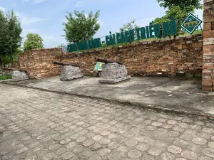 Hình ảnh chụp một khu trưng bày ngoài trời tại một di tích lịch sử. Trên một bức tường gạch đá thấp có dòng chữ lớn màu xanh lá cây ghi 'SÚNG THẦN CÔNG - CẢI CÁCH TRIỀU HỒ'. Bên phải có một biển báo hình thoi với biểu tượng cổng thành và dòng chữ 'THE HO CITADEL', 'THÀNH NHÀ HỒ', 'DI SẢN VĂN HÓA THẾ GIỚI'. Phía trước bức tường là ba khẩu súng thần công cổ, mỗi khẩu được đặt trên một bệ gạch riêng. Nền sân được lát bằng gạch khối. Phía sau là cây cối và bầu trời xanh có mây. Không có người hay hoạt động nào đang diễn ra trong ảnh.。（日本語訳）。ある史跡の屋外展示場を撮影した写真。低い石レンガの壁には、「SÚNG THẦN CÔNG - CẢI CÁCH TRIỀU HỒ」と記された緑色の大きな文字がある。右側には、城門のシンボルと「THE HO CITADEL」、「THÀNH NHÀ HỒ」、「DI SẢN VĂN HÓA THẾ GIỚI」という文字が入った菱形の標識がある。広場の地面はブロックレンガで敷き詰められている。後方には木々と、雲のある青空がある。写真には、写っている人や行われている活動はない。