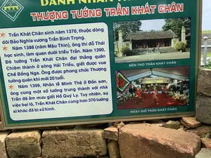 Đây là hình ảnh chụp cận cảnh một bảng thông tin tại một khu di tích lịch sử, có thể là Thành nhà Hồ dựa trên logo 'THE HO CITADEL' và 'THÀNH NHÀ HỒ DI SẢN VĂN HÓA THẾ GIỚI'. Bảng thông tin được đặt trên một bức tường thấp làm bằng những viên đá hoặc gạch cổ.。Nội dung văn bản trên bảng được ghi lại như sau:。Tiêu đề: 'DANH NHÂN THƯỢNG TƯỚNG TRẦN KHÁT CHÂN'.。Nội dung chính: 'Trần Khát Chân sinh năm 1370, thuộc dòng dõi Bảo Nghĩa vương Trần Bình Trọng. Năm 1388 (năm Mậu Thìn), ông thi đỗ Thái học sinh, làm quan dưới triều Trần. Năm 1390, Đô tướng Trần Khát Chân đại thắng quân Chiêm thành ở sông Hải Triều, giết được vua Chế Bồng Nga. Ông được phong chức Thượng tướng quân khi mới 20 tuổi. Năm 1399, Nhân lễ Minh Thệ ở Đốn sơn, ông cùng một số tướng trung thành với nhà Trần đã âm mưu giết Hồ Quý Ly. Tuy nhiên, sự việc bại lộ, Trần Khát Chân cùng hơn 370 tướng sĩ khác đã bị xử hình.'。Trên bảng có hai hình ảnh minh họa:。1. Một bức ảnh có chú thích 'ĐỀN THỜ TRẦN KHÁT CHÂN', cho thấy một công trình kiến trúc cổ với mái ngói, sân rộng và lư hương bằng đá.。2. Một bức ảnh có chú thích 'NGÀY GIỖ TRẦN KHÁT CHÂN', mô tả một hoạt động lễ hội hoặc nghi lễ tưởng niệm. Trong ảnh, nhiều người mặc trang phục truyền thống màu đỏ và vàng, cầm cờ hội, đang biểu diễn dưới một mái che lớn.。（日本語訳）。これは、歴史的遺跡にある案内板のクローズアップ写真です。「THE HO CITADEL」と「THÀNH NHÀ HỒ DI SẢN VĂN HÓA THẾ GIỚI」（胡朝の城塞 世界文化遺産）というロゴに基づくと、胡朝の城塞である可能性があります。案内板は、古い石またはレンガで作られた低い壁の上に設置されています。案内板のテキスト内容は以下の通りです。見出し：「名人 上将 陳渇真（チャン・カット・チャン）」。本文：「陳渇真は1370年に生まれ、保義王陳平仲（チャン・ビン・チョン）の子孫です。1388年（戊辰年）、彼は太学生の試験に合格し、陳朝の下で官吏となりました。1390年、都将陳渇真は海潮（ハイチェウ）川でチャンパ軍に大勝し、王である制蓬峨（チェー・ボン・ガー）を討ち取りました。彼はわずか20歳で上将軍の職に任命されました。1399年、頓山（ドンソン）での明誓の儀式の際、彼は陳朝に忠実な一部の将軍と共に胡季犛（ホー・クイ・リ）の暗殺を企てました。しかし、事が露見し、陳渇真は他の370人以上の将兵と共に処刑されました。」。案内板には2枚のイラスト写真があります。1. 「ĐỀN THỜ TRẦN KHÁT CHÂN」（陳渇真の祠）というキャプションが付いた写真で、瓦屋根、広い庭、石の香炉を持つ古い建築物を示しています。2. 「NGÀY GIỖ TRẦN KHÁT CHÂN」（陳渇真の命日）というキャプションが付いた写真で、祭りまたは追悼儀式の様子を描写しています。写真では、多くの人々が赤と黄色の伝統的な衣装を身に着け、祭りの旗を持ち、大きな屋根の下で演舞しています。