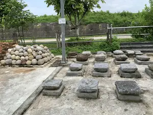 Đây là một khu vực trưng bày ngoài trời tại một di tích lịch sử, có thể là Thành nhà Hồ. Trên nền bê tông, nhiều khối đá lớn, có thể là chân tảng cột, được đặt trên các bệ xi măng. Một số khối đá này có chạm khắc hoa văn hình tròn trên bề mặt. Về phía bên trái, có một đống lớn các viên đá hình cầu, trông giống như đạn đá dùng cho máy bắn đá thời xưa, được xếp chồng lên nhau. Phía sau là một bức tường thành cổ được xây bằng những khối đá lớn, xung quanh có cây cối và thảm thực vật rậm rạp. Có một cột điện bằng kim loại, trên đó có một biển báo với nội dung: 'ĐIỆN CAO ÁP - TRÁNH XA', kèm theo dòng chữ tiếng Anh 'Dangerous Voltage - Keep Out'.。（日本語訳）。これは歴史的遺跡の屋外展示エリアで、おそらく胡朝の城塞でしょう。コンクリートの地面の上には、おそらく柱の礎石と思われる多くの大きな石のブロックが、セメントの台座の上に置かれています。これらの石のブロックの中には、表面に円形の模様が彫られているものもあります。後ろには大きな石のブロックで建てられた古い城壁があり、その周りには木々や鬱蒼とした植物が生い茂っています。金属製の電柱があり、そこには「ĐIỆN CAO ÁP - TRÁNH XA」（高圧電力 - 離れろ）という内容の看板があり、英語の「Dangerous Voltage - Keep Out」という文字が添えられています。