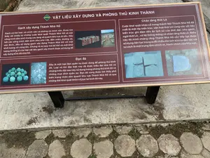 Bức ảnh chụp một bảng thông tin đặt ngoài trời tại một khu di tích lịch sử, có thể là Thành Nhà Hồ. Bảng có nền màu nâu, chữ trắng, được đặt trên hai cột đỡ màu đen trên nền xi măng và gạch lát. Bảng cung cấp thông tin về các vật liệu xây dựng và phòng thủ của kinh thành.。Nội dung văn bản trên bảng được ghi lại đầy đủ như sau:。Tiêu đề: VẬT LIỆU XÂY DỰNG VÀ PHÒNG THỦ KINH THÀNH。Nội dung bên trái:。- Gạch xây dựng Thành Nhà Hồ: 'Gạch có hai loại: có minh văn và không có minh văn. Được tìm thấy rất nhiều từ những cuộc khai quật Thành Nhà Hồ và các công trình dân sinh trong các làng xã quanh khu vực di sản. Hầu hết các công trình dân sinh như nhà cửa, đền miếu, đường xá, sân đình...đều sử dụng gạch xây dựng Thành Nhà Hồ với một khối lượng vô cùng lớn. Chứng tỏ sự quy mô bề thế và một khối lượng khổng lồ của loại vật liệu gạch xây thành được sử dụng để xây dựng kinh đô nhà Hồ.'。- Đạn đá: 'Đây là một loại đạn quân sự được dùng để phòng thủ kinh đô. Loại vũ khí đặc biệt này đã được triều đại nhà Hồ và những triều đại sau này sử dụng trong phòng thủ kinh đô và những mục đích quân sự. Đạn đá cũng được tìm thấy phổ biến trong nhân dân quanh khu vực Thành Nhà Hồ và qua những đợt khai quật khảo cổ học tại kinh đô này.'。Nội dung bên phải:。- Chân tảng thời Lê: 'Cuộc khai quật khảo cổ trong thành Nội Thành Nhà Hồ đã xuất lộ một lớp kiến trúc, văn hoá thời Lê cùng các vật liệu kiến trúc ghi đậm dấu ấn lịch sử của thời đại: như gạch, ngói thời Lê... và hệ thống móng và chân tảng thời Lê. Điều này đã minh chứng rằng Thành Nhà Hồ vẫn tiếp tục được sử dụng liên tục trong suốt chiều dài lịch sử của dân tộc với tư cách là một trung tâm chính trị, hành chính của khu vực.'。Các hình ảnh trên bảng minh họa cho nội dung, bao gồm: gạch cổ, khu vực khai quật khảo cổ, đạn đá và các khối chân tảng bằng đá.。（日本語訳）。写真は、歴史的遺跡、おそらく胡朝の城塞に屋外に設置された案内板を写したものです。板は茶色の背景に白文字で、セメントと敷石の土台の上にある2本の黒い支柱に設置されています。板は、都城の建築材料と防御に関する情報を提供しています。板に書かれた文章の全文は以下の通りです。見出し：都城の建築材料と防御。左側の内容：。- 胡朝の城塞の建築用レンガ：「レンガには2種類あります：銘文があるものと銘文がないものです。胡朝の城塞の発掘調査や、遺産地域周辺の村々の民間施設から多数発見されています。家屋、祠堂、道路、村の集会所の庭など、ほとんどの民間施設は、胡朝の城塞の建築用レンガを非常に大量に使用しています。これは、胡朝の都を建設するために使用された城壁用レンガという材料の壮大な規模と膨大な量を示しています。」。- 石弾：「これは都城を防御するために使用された軍事用の弾丸の一種です。この特殊な武器は、胡朝とその後の王朝によって、都城の防御や軍事目的で使用されました。石弾は、胡朝の城塞周辺の住民の間でも、またこの都城での考古学的な発掘調査を通じても、広く発見されています。」。右側の内容：。- 黎朝時代の礎石：「胡朝の城塞の内城における考古学的な発掘調査により、黎朝時代の建築、文化の層が、その時代の歴史的痕跡を色濃く残す建築材料、例えば黎朝時代のレンガ、瓦…そして黎朝時代の基礎と礎石のシステムと共に出土しました。このことは、胡朝の城塞が、地域の政治・行政の中心として、民族の歴史の長きにわたって継続的に使用され続けたことを証明しています。」。板上の写真は内容を説明するもので、古いレンガ、考古学発掘区域、石弾、石の礎石ブロックなどが含まれています。