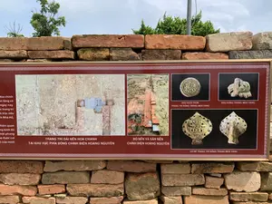 Bức ảnh chụp một tấm bảng thông tin được đặt ngoài trời tại một khu di tích lịch sử, phía trước một bức tường xây bằng gạch cổ. Tấm bảng có viền màu đỏ với họa tiết trang trí, bên trong hiển thị các hình ảnh và chú thích về những phát hiện khảo cổ học.。Nội dung văn bản trên bảng:。- Một cột văn bản bên trái bị che khuất một phần, chỉ có thể đọc được các cụm từ như: "...tòa chính", "...ngày nay.", "...trang trí", "...h Nhà Hồ,", "...hiện quan", "...ng kinh đô".。- Chú thích cho các hình ảnh:。+ "TRANG TRÍ DẢI NỀN HOA CHANH TẠI KHU VỰC PHÍA ĐÔNG CHÍNH ĐIỆN HOÀNG NGUYÊN"。+ "BÓ NỀN VÀ SÂN NỀN CHÍNH ĐIỆN HOÀNG NGUYÊN"。+ "ĐẦU NGÓI TRÒN TRANG TRÍ RỒNG TRÁNG MEN VÀNG"。+ "ĐẦU RỒNG TRÁNG MEN VÀNG"。+ "LÁ ĐỀ TRANG TRÍ RỒNG TRÁNG MEN VÀNG"。Cảnh vật và hoạt động:。- Địa điểm: Một khu di tích khảo cổ, có thể là Thành nhà Hồ dựa trên nội dung văn bản.。- Hoạt động: Không có hoạt động nào của con người diễn ra. Bức ảnh ghi lại một hiện trạng tĩnh của tấm bảng thông tin.。- Các vật thể được chụp: Tấm bảng thông tin, tường gạch cổ, cây cối và bầu trời. Các hình ảnh trên bảng mô tả các hiện vật khảo cổ như nền móng công trình, đầu ngói, đầu rồng và lá đề trang trí bằng gốm tráng men vàng.。（日本語訳）。写真は、歴史遺跡の屋外に設置された案内板を撮影したもので、古いレンガで造られた壁の前にあります。案内板は装飾模様のある赤い縁取りで、内部には考古学的発見に関する画像と説明が表示されています。案内板のテキスト内容：。- 左側のテキスト欄は一部が隠れており、「...正殿」、「...今日。」、「...装飾」、「...胡朝の城」、「...現官」、「...都」などの語句しか読み取れません。- 画像の説明：。+ 「皇元正殿東側区域におけるライムの花模様の基礎装飾」。+ 「皇元正殿の基礎の縁と床」。+ 「黄釉龍装飾の丸瓦頭」。+ 「黄釉の龍の頭」。+ 「黄釉龍装飾の菩提樹の葉」。風景と活動：。- 場所：考古学遺跡。テキスト内容に基づくと、胡朝の城である可能性があります。- 活動：人間の活動は見られません。写真は案内板の静的な状態を記録しています。- 撮影された物体：案内板、古いレンガの壁、木々、空。案内板の画像は、建物の基礎、瓦頭、龍の頭、黄釉陶器で装飾された菩提樹の葉などの考古学的遺物を描写しています。