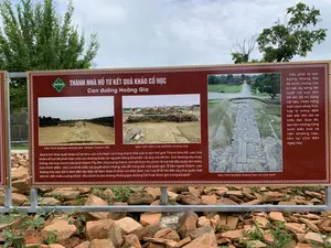 Đây là một bảng thông tin được đặt ngoài trời tại một địa điểm khảo cổ, cụ thể là khu di sản Thành Nhà Hồ. Bảng có nền màu nâu, chữ trắng và viền hoa văn trang trí.。**Nội dung chữ viết trên bảng:**。**Tiêu đề chính:** THÀNH NHÀ HỒ TỪ KẾT QUẢ KHẢO CỔ HỌC - Con đường Hoàng Gia。**Nội dung bên trái:**。'Quá trình khai quật khảo cổ tại khu vực cửa Nam và trong thành Nội của di sản thế giới Thành Nhà Hồ, các nhà khảo cổ học đã phát hiện một con đường được lát nguyên bằng đá phiến với quy mô rất lớn. Con đường này chạy thẳng vào trục chính của kinh thành Tây Đô. Vào trong thành, khi kết thúc thì chính là nơi bắt đầu bước lên thềm của chính điện - nơi vua thiết triều và quyết định những vấn đề trọng đại của quốc gia. Ở bên ngoài thành, con đường này kéo dài 2.5km đến tận Đàn tế Nam Giao ở chân núi Đốn Sơn- nơi vua tế lễ trời đất cầu cho quốc thái dân an, đất nước cường thịnh - Đó chính là con đường Hoàng gia (đường Cái hoa) được ghi trong sử sách.'。**Nội dung bên phải:**。'Việc phát lộ con đường Hoàng Gia đã minh chứng cho trí tuệ, sự sáng tạo tuyệt vời của nhà Hồ khi đã sử dụng vật liệu, nhân công một cách khoa học, hợp lý mang đậm bản sắc dân tộc và triều đại. Qua đó, tạo nên những công trình kiến trúc vô tiền khoáng hậu, tồn tại cho đến ngày nay.'。**Chú thích ảnh:**。- DẤU TÍCH ĐƯỜNG HOÀNG GIA TRONG THÀNH NỘI。- MẶT CẮT NỀN CON ĐƯỜNG HOÀNG GIA。- DẤU TÍCH ĐƯỜNG HOÀNG GIA TẠI CỬA NAM。**Mô tả quang cảnh và vật thể:**。- **Địa điểm:** Khu di tích khảo cổ ngoài trời, thuộc Thành Nhà Hồ.。- **Hoạt động:** Bảng thông tin đang thực hiện chức năng cung cấp thông tin lịch sử và khảo cổ học cho khách tham quan. Không có hoạt động của con người trong ảnh.。- **Vật thể:**。- Bảng thông tin lớn bằng kim loại.。- Các hình ảnh chụp lại quá trình khai quật và di tích con đường đá.。- Phía dưới bảng là các mảnh gạch và đá vỡ, có thể là một phần của di tích được trưng bày.。- Phía sau là cây cối xanh tươi và bầu trời có mây.。（日本語訳）。これは、考古学的な場所、具体的には胡朝の城塞遺跡に屋外に設置された案内板です。板は茶色の背景に白い文字で、装飾的な模様の縁取りがあります。**板に書かれた内容：**。**主な見出し：** 考古学的成果から見る胡朝の城塞 - 王の道。**左側の内容：**。「世界遺産である胡朝の城塞の南門区域と内城での考古学的発掘調査の過程で、考古学者たちは非常に大規模な、完全にスレート石で舗装された道を発見しました。この道は西都（タイドー）の都城の主軸にまっすぐ続いています。城内に入ると、その終点は正殿の階段の始まりであり、そこは王が朝廷を開き、国家の重大な問題を決定する場所でした。城外では、この道はドンソン山の麓にある南郊壇まで2.5km伸びており、そこは王が天地を祀り、国泰民安と国家の繁栄を祈る場所でした。これこそが、史書に記されている王の道（カイホア通り）です。」。**右側の内容：**。「王の道の発見は、胡朝が資材と労働力を科学的かつ合理的に使用し、民族と王朝の特色を色濃く反映させた、その知恵と素晴らしい創造性を証明しました。それにより、今日まで存在する空前絶後の建築物を生み出したのです。」。**写真の説明：**。- 内城にある王の道の痕跡。- 王の道の基盤断面。- 南門にある王の道の痕跡。**景観と物体の説明：**。- **場所：** 胡朝の城塞に属する屋外の考古学遺跡。- **活動：** 案内板は、見学者に歴史的および考古学的な情報を提供する機能を果たしています。写真には人間の活動はありません。- **物体：**。- 金属製の大きな案内板。- 発掘過程と石の道の遺跡を撮影した写真。- 板の下には、展示されている遺跡の一部である可能性のある、割れたレンガや石の破片があります。- 後ろには青々とした木々と雲のある空があります。