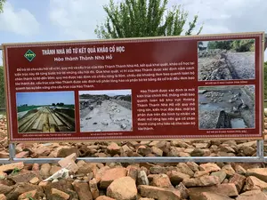 Đây là hình ảnh một tấm bảng thông tin lớn được đặt ngoài trời tại một di tích lịch sử, có thể là khu vực Thành Nhà Hồ. Tấm bảng có nền màu nâu, chữ màu trắng, được đặt trên một đống đá và gạch vỡ. Phía sau là cây cối và bầu trời.。Nội dung văn bản trên bảng như sau:。Tiêu đề chính: THÀNH NHÀ HỒ TỪ KẾT QUẢ KHẢO CỔ HỌC。Tiêu đề phụ: Hào thành Thành Nhà Hồ。Nội dung chi tiết:。'Để trả lời cho câu hỏi về vị trí, quy mô và cấu trúc của Hào Thành Thành Nhà Hồ, kết quả khai quật, khảo cổ học tại kiến trúc này đã từng bước trả lời những câu hỏi đó. Qua khai quật, vị trí của Hào Thành được xác định nằm cách chân thành từ 60 đến 90m; quy mô được xác định với chiều rộng là 50m, chiều dài khoảng 4km bao quanh toàn bộ tòa thành đá; cấu trúc của Hào Thành được xác định với phần lòng hào và phần bờ kè bằng đá cổ trải đều 4km bao quanh tạo sự bền vững cho cấu trúc của Hào Thành.'。'Hào Thành được xác định là một kiến trúc chỉnh thể, thống nhất bao quanh toàn bộ khu vực Hoàng Thành Thành Nhà Hồ với quy mô rộng lớn, kiến trúc độc đáo, một bộ phận dựa trên địa hình tự nhiên và được mở rộng tạo nền gia cố chân thành cũng như bảo vệ cho toàn bộ tòa thành.'。Bảng còn có bốn bức ảnh chụp các địa điểm khai quật khảo cổ, minh họa cho các hoạt động nghiên cứu. Các chú thích cho ảnh bao gồm:。- CHIỀU RỘNG HÀO NAM VÀ DẤU TÍCH ĐÁ GIA CỐ BỜ HÀO。- BỜ KÈ ĐÁ Ở HÀO THÀNH PHÍA TÂY。- BỜ KÈ ĐÁ Ở HÀO THÀNH PHÍA ĐÔNG。- BỜ KÈ ĐÁ Ở HÀO THÀNH PHÍA BẮC。Các bức ảnh cho thấy các hố và rãnh khai quật, để lộ các lớp đất và cấu trúc đá của hào thành cổ. Một số ảnh có hình người đang làm việc tại công trường khảo cổ.。（日本語訳）。これは、史跡、おそらく胡朝の城塞のエリアに屋外に設置された大きな案内板の写真です。案内板は茶色の背景に白い文字で、石と割れたレンガの山の上に置かれています。後ろには木々と空が見えます。板に書かれている文章の内容は以下の通りです。主な見出し：考古学的成果から見た胡朝の城塞。副見出し：胡朝の城塞の堀。詳細内容：。「胡朝の城塞の堀の位置、規模、構造に関する問いに答えるため、この建造物における発掘調査、考古学の成果が、それらの問いに一歩ずつ答えてきました。発掘調査を通じて、堀の位置は城壁の麓から60～90mの距離にあると特定されました。規模は幅50m、長さ約4kmで石の城全体を囲んでいると特定されました。堀の構造は、堀底部分と古代の石でできた護岸部分が4kmにわたって均等に広がり、堀の構造の持続性を生み出していると特定されました。」。「堀は、胡朝の城塞の皇城エリア全体を囲む、一体的で統一された建造物であり、広大な規模と独特の建築様式を持ち、一部は自然の地形に基づいており、城壁の麓を補強し、城全体を保護するために拡張されたと特定されています。」。板には、研究活動を説明するために、考古学的な発掘現場を撮影した4枚の写真もあります。写真のキャプションには以下が含まれます。- 南堀の幅と堀岸を補強した石の痕跡。- 西側の堀の石積み護岸。- 東側の堀の石積み護岸。- 北側の堀の石積み護岸。写真には、発掘された穴や溝が写っており、古代の堀の土層や石の構造が露出しています。