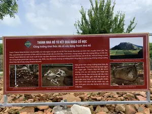 Bức ảnh chụp một tấm biển thông tin lớn được đặt ngoài trời tại một địa điểm khảo cổ học gần Thành Nhà Hồ. Tấm biển có nền màu nâu đỏ, chữ trắng, và khung viền hoa văn, được dựng trên một khung kim loại trước một đống đá. Phía sau là cây cối và bầu trời.。Nội dung văn bản trên biển được ghi lại đầy đủ như sau:。Tiêu đề chính: THÀNH NHÀ HỒ TỪ KẾT QUẢ KHẢO CỔ HỌC。Tiêu đề phụ: Công trường khai thác đá cổ xây dựng Thành Nhà Hồ。Nội dung bên trái: "Cách Thành Nhà Hồ 2km về phía Bắc có một ngọn núi gọi là núi An Tôn thuộc địa phận xã Vĩnh Yên, huyện Vĩnh Lộc. Tại đây, qua khảo sát và khảo cổ đã xuất lộ nhiều phiến đá có chất liệu, hình thức, trọng lượng, kết cấu tương đồng với đá xây dựng Thành Nhà Hồ, bên dưới xuất lộ các lớp dăm đá được hình thành từ quá trình chế tác các khối đá, một số công cụ chế tác cũng được phát hiện tại khu vực này. Điều này đã minh chứng và khẳng định rằng đây là một trong những công trường khai thác đá cổ xây dựng Thành Nhà Hồ."。Nội dung bên phải: "Việc phát hiện công trường khai thác đá cổ xây dựng Thành Nhà Hồ tại một ngọn núi ngay gần Thành Nhà Hồ đã minh chứng cho sự vận dụng linh hoạt của Nhà Hồ trong việc sử dụng vật liệu hiện có và ở địa điểm gần nhất để thuận lợi cho việc khai thác và vận chuyển nhằm tiết kiệm một cách tối đa nhân lực, vật lực, thời gian cho xây dựng kinh đô của triều đại."。Trên biển có bốn hình ảnh minh họa kèm chú thích:。1.  "TOÀN CẢNH NÚI AN TÔN - CÔNG TRƯỜNG KHAI THÁC ĐÁ CỔ": Hình ảnh một ngọn núi với các thửa ruộng bậc thang hoặc cánh đồng ở chân núi.。2.  "LỚP DĂM ĐÁ TẠI CÔNG TRƯỜNG KHAI THÁC ĐÁ CỔ": Hình ảnh một lớp đất chứa đầy đá vụn (dăm đá) và một cây thước đo khảo cổ học.。3.  "KHAI QUẬT TẠI CÔNG TRƯỜNG KHAI THÁC ĐÁ CỔ": Hình ảnh một khu vực khai quật với nhiều khối đá lớn nhỏ nằm trên mặt đất.。4.  "PHIẾN ĐÁ TẠI CÔNG TRƯỜNG ĐÁ CỔ": Hình ảnh một khối đá lớn, hình chữ nhật, có dấu vết của việc đẽo gọt.。（日本語訳）。胡朝の城塞近くの考古学遺跡に屋外設置された、大きな案内板の写真。案内板は赤褐色の背景に白文字、模様の縁取りがあり、石の山の前に金属製のフレームで立てられている。案内板に書かれている文章の全文は以下の通りである。主題：考古学的成果から見た胡朝の城塞。副題：胡朝の城塞建設のための古代石材採掘場。左側の内容：「胡朝の城塞から北へ2km、ヴィンロック県ヴィンイエン社に属するアントン山という山があります。ここでは、調査と考古学的発掘を通じて、胡朝の城塞の建設に使用された石材と材質、形状、重量、構造が類似する多くの石板が露出し、その下からは石材を加工する過程で生じた砕石の層が露出し、この地域ではいくつかの加工道具も発見されました。このことは、ここが胡朝の城塞を建設するための古代石材採掘場の一つであったことを証明し、断定するものです。」。右側の内容：「胡朝の城塞のすぐ近くの山で、城塞建設のための古代石材採掘場が発見されたことは、胡朝が既存の材料を最も近い場所で利用し、採掘と輸送を容易にすることで、王朝の首都建設のための人的資源、物的資源、時間を最大限に節約するという、柔軟な応用力を証明するものです。」。案内板には、キャプション付きの4枚のイラスト写真がある。1. 「アントン山の全景 - 古代石材採掘場」：山の麓に棚田または畑が広がる山の写真。2. 「古代石材採掘場の砕石層」：砕石（チップ）で満たされた土層と考古学用の物差しが写った写真。3. 「古代石材採掘場の発掘」：大小さまざまな石塊が地面に転がっている発掘エリアの写真。4. 「古代石材採掘場の石板」：削られた跡のある、大きな長方形の石塊の写真。