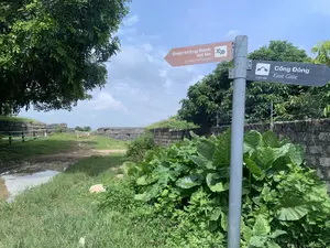 Hình ảnh chụp tại một khu di tích lịch sử ngoài trời, có một cột biển chỉ dẫn bằng kim loại. Trên cột có hai biển báo chỉ hướng cho khách tham quan:。- Biển báo màu nâu chỉ về bên trái có ghi: "Đoạn tường thành sụt lún" và "Sunken Wall Section".。- Biển báo màu đen chỉ về bên phải có ghi: "Cổng Đông", "East Gate", và khoảng cách "130 m".。Bối cảnh là một con đường đất có vũng nước, chạy dọc theo một bức tường thành cổ được xây bằng những khối đá lớn. Xung quanh có nhiều cây cối, cỏ và các bụi cây lá to mọc um tùm. Bầu trời trong xanh, có mây trắng. Không có người hay hoạt động nào được ghi lại trong ảnh.。（日本語訳）。屋外の史跡で撮影された写真で、金属製の案内標識柱が一本あります。柱には、観光客向けの方向を示す2つの標識があります：。- 左を指す茶色の標識には、「Đoạn tường thành sụt lún」と「Sunken Wall Section」と書かれています。- 右を指す黒色の標識には、「Cổng Đông」、「East Gate」、そして距離「130 m」と書かれています。背景は、大きな石のブロックで築かれた古い城壁に沿って続く、水たまりのある未舗装の道です。周囲には多くの木々、草、そして大きな葉を持つ茂みが鬱蒼と生い茂っています。空は青く澄んでおり、白い雲があります。写真には人や活動は記録されていません。