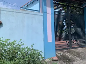 Hình ảnh cho thấy một phần bên ngoài của một ngôi nhà. Bên trái là một bức tường được sơn màu xanh da trời. Bên cạnh bức tường là một cột cổng được sơn màu xanh lam và hồng nhạt. Cánh cổng chính được làm bằng sắt màu đen, có các thanh dọc và ngang, với một họa tiết trang trí hình tròn ở giữa. Nhìn qua cổng, có thể thấy khoảng sân được lát gạch màu nâu đỏ. Bên trong sân có một chiếc xe đạp đang đỗ. Ngôi nhà phía sau cũng được sơn màu xanh, có mái lợp ngói đỏ. Trên tường nhà, phía sau cổng, có một biểu tượng hình tròn màu vàng có chữ nhưng không thể đọc rõ. Phía trước bức tường bên trái có một bụi cây xanh. Bầu trời trong xanh, cho thấy đây là một cảnh ban ngày.。（日本語訳）。画像は家の一部分の外観を示しています。左側には水色に塗られた壁があります。壁の隣には、青色と薄いピンク色に塗られた門柱があります。正門は黒い鉄でできており、縦と横の格子があり、中央には円形の装飾模様があります。門を通して、赤茶色のタイルが敷かれた庭が見えます。庭の中には自転車が一台停まっています。奥の家も青色に塗られており、赤い瓦屋根があります。左側の壁の前には緑の茂みがあります。空は青く澄んでおり、昼間の光景であることがわかります。