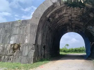 Hình ảnh chụp một cổng thành cổ bằng đá rất lớn và kiên cố, có kiến trúc hình vòm. Bức tường thành và cổng được xây dựng từ những phiến đá lớn, đã ngả màu cũ kỹ và có dấu hiệu của sự phong hóa theo thời gian, với một ít cây cỏ mọc trên đỉnh và các kẽ đá. Một con đường đất sỏi nhỏ dẫn xuyên qua cổng vòm. Nhìn qua vòm cổng, có thể thấy khung cảnh vùng nông thôn với cây cối xanh tươi, một cây cao đơn độc và bầu trời trong xanh với vài gợn mây. Không có người hay hoạt động nào diễn ra. Không có văn tự hay chữ viết nào có thể nhận dạng được trên các phiến đá. Đây là một di tích lịch sử.。（日本語訳）。非常に大きく頑丈な石造りの古い城門の写真で、アーチ状の建築様式をしている。城壁と門は大きな石板から造られており、古びた色に変色し、経年による風化の兆候が見られ、頂上や石の隙間には少量の草木が生えている。小さな砂利道がアーチ門を通り抜けている。人や活動は見られない。石板には識別できる文字や記述はない。これは歴史的な遺跡である。