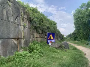 Bản ghi văn bản trên biển báo: 'KHU VỰC SẠT LỞ NGUY HIỂM. ĐỀ NGHỊ KHÔNG DỪNG LẠI'. Một phần chữ ở dòng dưới cùng bị che khuất, chỉ có thể đọc được chữ 'THAM'.。Bối cảnh là một khu vực ngoài trời, có một bức tường thành cổ rất lớn được xây dựng từ những khối đá khổng lồ, xếp chồng lên nhau. Bức tường này có dấu hiệu của thời gian và bị cây cỏ dại mọc um tùm trên đỉnh và len lỏi qua các kẽ đá. Bên cạnh bức tường là một con đường đất nhỏ. Ở phía trước, có một biển báo nguy hiểm màu xanh lam với biểu tượng hình tam giác màu vàng cảnh báo về nguy cơ sạt lở đất đá. Không có người trong ảnh. Khung cảnh xung quanh chủ yếu là cây xanh, cỏ dại, và một vài tảng đá lớn nằm trên mặt đất. Phía trên có thể thấy dây điện vắt ngang qua bầu trời xanh và có mây.。（日本語訳）。看板のテキスト記録：「危険な地滑り区域。停止しないようお願いします」。最下行の文字の一部が隠れており、「THAM」という文字しか読めない。背景は屋外のエリアで、巨大な石のブロックを積み重ねて作られた非常に大きな古い城壁がある。壁の隣には小さな未舗装の道がある。前面には、土砂崩れの危険を警告する黄色の三角形のシンボルが付いた青い危険標識がある。写真には人がいない。周囲の風景は主に緑の木々、雑草、そして地面に横たわるいくつかの大きな岩である。