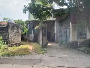 Quang cảnh một con ngõ nhỏ tại một khu dân cư ở vùng nông thôn. Bên trái là một lối đi nhỏ bằng đất bên cạnh một bức tường gạch cũ. Bên phải là lối vào một ngôi nhà có sân bê tông và cổng sắt màu xám. Ngôi nhà này có tường rào bê tông với hàng rào sắt màu xanh lá cây, trên đó có giàn hoa giấy. Phía sau là một ngôi nhà nhiều tầng sơn màu xanh lá cây. Cây cối xanh tươi mọc xung quanh. Trên một mái nhà có một bồn chứa nước bằng inox. Có thể thấy một chiếc xe đạp dựng trong sân. Trên các cột cổng bê tông có ghi các con số, bao gồm '3560' ở cột bên trái và '0344... 7374' ở cột bên phải, một số chữ số bị mờ không đọc được. Khung cảnh tĩnh lặng, không có người hay hoạt động nào đang diễn ra.。（日本語訳）。田舎の住宅地にある小さな路地の光景。左側は、古いレンガの壁の隣にある小さな土の道です。右側は、コンクリートの庭と灰色の鉄の門がある家への入り口です。この家には緑色の鉄柵が付いたコンクリートの塀があり、その上にはブーゲンビリアの棚があります。奥には、緑色に塗られた多層階の家があります。周りには青々とした木々が生い茂っています。ある屋根の上にはステンレス製の貯水タンクがあります。庭に立てかけられた自転車が見えます。コンクリートの門柱には数字が書かれており、左の柱には「3560」、右の柱には「0344... 7374」とありますが、一部の数字はかすれていて読めません。静かな光景で、人や進行中の活動はありません。