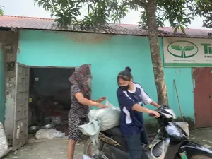 Bối cảnh diễn ra ở phía trước một ngôi nhà cấp bốn ở vùng nông thôn, có bức tường được sơn màu xanh ngọc và mái lợp tôn. Có hai người phụ nữ đang chất một bao tải lớn màu trắng lên yên sau của một chiếc xe máy. Một người phụ nữ lớn tuổi hơn, đội mũ chống nắng có hoa văn, đang phụ giúp. Người còn lại là một cô gái trẻ, đeo khẩu trang, đang ngồi trên xe máy và giữ tay lái, chuẩn bị vận chuyển bao hàng. Nhìn vào trong nhà qua ô cửa mở, có thể thấy một vài chiếc chậu nhôm và các bao tải khác, có một ít khói bốc ra từ bên trong. Trên tường bên ngoài có treo một tấm biển hiệu với logo hai chiếc lá màu xanh và dòng chữ 'WWW.TIENNONG.VN'. Phần chữ lớn trên biển hiệu có thể đọc được một phần là 'TI... DINH...'.。（日本語訳）。舞台は田舎にある平屋建ての家の前で、壁はエメラルドグリーンに塗られ、屋根はトタンで葺かれている。二人の女性が、大きな白い袋をバイクの後部座席に積んでいる。年配の女性は、花柄の日よけ帽子をかぶり、手伝っている。もう一人は若い女性で、マスクを着け、バイクにまたがってハンドルを握り、荷物を運ぶ準備をしている。開いた戸口から家の中を覗くと、いくつかのアルミ製の鉢や他の袋が見え、中から少し煙が立ち上っている。看板の大きな文字は、「TI... DINH...」と一部読み取れる。