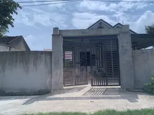 Bức ảnh chụp cổng vào của một ngôi nhà ở một vùng có vẻ là nông thôn hoặc ngoại ô tại Việt Nam. Cổng chính được xây dựng kiên cố với hai cột bê tông lớn và một bộ cửa sắt màu sẫm, thiết kế hiện đại. Một cánh cửa đang mở hé, cho thấy lối vào sân trong. Qua cánh cổng, có thể nhìn thấy một phần của ngôi nhà với mái ngói kiểu mới. Bên trái cổng là một bức tường bê tông cũ kỹ. Phía trên là bầu trời xanh có mây trắng và các đường dây điện, dây cáp vắt ngang. Khung cảnh tĩnh lặng, không có hoạt động nào của con người diễn ra.。（日本語訳）。ベトナムの田舎か郊外と思われる地域にある家の入り口の門を撮影した写真。正門は、2本の大きなコンクリートの柱と、モダンなデザインの暗い色の鉄の扉で頑丈に建てられている。門の左側には、古びたコンクリートの壁がある。上には、白い雲のある青空と、横切る電線やケーブルがある。静かな光景で、人の活動は一切行われていない。