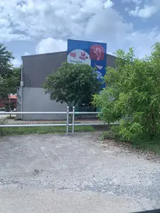 Chữ viết được ghi lại: Trên biển hiệu lớn màu xanh dương có chữ "Trưng bày". Trên cửa ra vào có chữ "LỐI VÀO" và "ENTRANCE".。Mô tả cảnh vật và hoạt động: Bức ảnh chụp bên ngoài một tòa nhà lớn, có kiến trúc hiện đại giống nhà xưởng với tường bê tông và phần trên bằng tôn lượn sóng màu xám. Dựa vào tấm biển hiệu, đây có thể là một nhà trưng bày hoặc trung tâm triển lãm, có khả năng thuộc về một khu di tích. Một tấm biển hiệu lớn màu xanh dương được gắn trên tường, có chữ "Trưng bày" và hình ảnh các hiện vật trông giống như đồ gốm hoặc các chi tiết kiến trúc cổ bằng đất nung. Phía trước tòa nhà có cây xanh và bụi rậm. Một hàng rào kim loại màu bạc nằm ở tiền cảnh. Lối đi phía trước được làm bằng bê tông và sỏi đá. Bầu trời trong ảnh có mây. Không có hoạt động nào của con người được ghi lại.。（日本語訳）。記録された文字：大きな青い看板に「Trưng bày」（展示）という文字がある。出入口のドアに「LỐI VÀO」（入口）と「ENTRANCE」という文字がある。風景と活動の説明：写真は大きな建物の外観を撮影したもので、コンクリートの壁と灰色の波板でできた上部を持つ、工場のような現代的な建築様式である。看板から判断すると、ここは展示館または展示センターであり、史跡の一部である可能性がある。建物の前には緑の木々と茂みがある。前景には銀色の金属製の柵がある。前面の通路はコンクリートと砂利でできている。写真の空は曇っている。人間の活動は記録されていない。