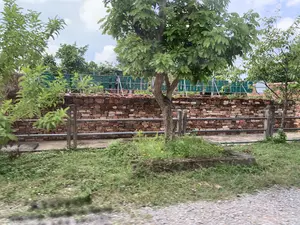 Trong ảnh là một khung cảnh ngoài trời, có thể là một phần của một di tích lịch sử. Nổi bật là một bức tường dài được xây bằng gạch cũ, có màu nâu đỏ. Phía trước bức tường là một hàng rào thấp làm bằng các cột bê tông và thanh chắn kim loại. Ở tiền cảnh là một con đường rải sỏi và một khu đất nhỏ có cỏ và một cái cây. Phía sau bức tường gạch, có một tấm biển lớn màu xanh lá cây với các chữ cái màu trắng, nhưng nội dung không thể đọc rõ do bị cây cối che khuất. Không có người hoặc hoạt động cụ thể nào được ghi lại trong ảnh.。（日本語訳）。写真には屋外の風景が写っており、おそらく史跡の一部だと思われます。目立っているのは、赤褐色の古いレンガで造られた長い壁です。壁の前には、コンクリートの柱と金属の柵でできた低いフェンスがあります。前景には砂利道と、草と一本の木が生えている小さな土地があります。レンガの壁の後ろには、白い文字が書かれた大きな緑色の看板がありますが、木々に隠れているため内容ははっきりと読み取れません。写真には、人物や特定の活動は写っていません。