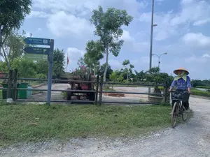 Nội dung trên biển chỉ dẫn:。- Biển màu xanh lam chỉ hướng đến "Nhà trưng bày (Exhibitions)", cách 140 m.。- Biển màu xanh đậm chỉ hướng đến "Bãi đỗ xe (Parking)", cách 150 m.。- Biển màu xanh lá cây chỉ hướng đến "Trung tâm Thông tin Du khách (Visitor Information Center)", cách 140 m.。Bối cảnh là một con đường sỏi đá ở khu vực nông thôn, có thể là lối vào của một khu du lịch hoặc di tích. Một người phụ nữ đội nón lá, mặc áo dài tay màu xanh, đang đi xe đạp trên đường. Bên vệ đường có một cột biển báo. Phía sau hàng rào là một khoảng sân rộng, nơi có một chiếc xe kéo bằng gỗ được trang trí bằng các chậu hoa. Xung quanh có cây cối, cột đèn và ở phía xa có một lá cờ đang bay. Bầu trời trong xanh và có mây trắng.。（日本語訳）。案内標識の内容：。- 水色の標識は「展示館（Exhibitions）」の方向を示しており、140m先です。- 濃い青色の標識は「駐車場（Parking）」の方向を示しており、150m先です。- 緑色の標識は「ビジターインフォメーションセンター（Visitor Information Center）」の方向を示しており、140m先です。背景は田舎の砂利道で、観光地または史跡の入り口である可能性があります。ノンラーをかぶり、青い長袖の服を着た女性が、道を自転車で走っています。道端には案内標識の柱が一本あります。柵の後ろには広い庭があり、そこには植木鉢で飾られた木製の荷車があります。周りには木々や街灯があり、遠くには旗がはためいています。空は青く澄んでおり、白い雲があります。