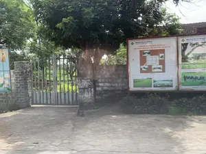 Bối cảnh là một khu vực ngoài trời, dường như là lối vào của khu di tích Thành Nhà Hồ.。**Chữ viết trên bảng thông tin:** Có các bảng thông tin lớn giới thiệu về di tích. Một bảng có tiêu đề nổi bật, mặc dù hơi mờ: 'THÀNH NHÀ HỒ VỚI GIÁ TRỊ NỔI BẬT TOÀN CẦU. ĐƯỢC UNESCO CÔNG NHẬN DI SẢN THẾ GIỚI...'. Phía trên cùng của bảng có ghi 'TRUNG TÂM BẢO TỒN DI SẢN THÀNH NHÀ HỒ (CONSERVATION CENTER FOR HO CITADEL)'. Trên mái nhà phía xa có một tấm biển nhỏ ghi số '19'. Ở bên trái, có một tấm biển nhỏ hơn treo trên cột với hình ảnh Đức Phật ngồi thiền và hoa sen, nhưng chữ viết trên đó không thể đọc rõ.。**Cảnh vật và sự vật:** Khung cảnh bao gồm một lối đi bằng bê tông dẫn đến một chiếc cổng sắt. Hai bên cổng là các cột tường được xây bằng gạch mộc. Các bảng thông tin lớn được dựng lên để cung cấp thông tin cho du khách, trên đó có hình ảnh về các công trình của di tích như cổng thành đá, sơ đồ khu vực và cảnh quan xung quanh. Phía sau cánh cổng sắt là một cánh đồng xanh mướt, có thể nhìn thấy một con vật (trông giống trâu hoặc bò). Xung quanh có nhiều cây xanh lớn. Không có hoạt động nào của con người diễn ra trong ảnh.。（日本語訳）。背景は屋外で、胡朝の城塞遺跡の入り口のようです。**案内板の文字：** 遺跡を紹介する大きな案内板があります。一つの案内板には、少しぼやけてはいますが、目立つ見出しがあります：「全世界的な顕著な価値を持つ胡朝の城。ユネスコにより世界遺産に認定…」。案内板の最上部には「TRUNG TÂM BẢO TỒN DI SẢN THÀNH NHÀ HỒ (CONSERVATION CENTER FOR HO CITADEL)」と書かれています。遠くの屋根の上には、「19」という数字が書かれた小さな看板があります。左側には、柱に掛けられた小さな看板があり、瞑想する仏陀と蓮の花の絵が描かれていますが、その上の文字ははっきりと読むことができません。**風景と物：** 風景には、鉄の門に通じるコンクリートの通路が含まれています。門の両側には、むき出しのレンガで建てられた壁の柱があります。観光客に情報を提供するために大きな案内板が設置されており、その上には石の城門などの遺跡の建造物、エリアの地図、周辺の景観の写真が掲載されています。周りには大きな緑の木がたくさんあります。写真の中では人間の活動は見られません。