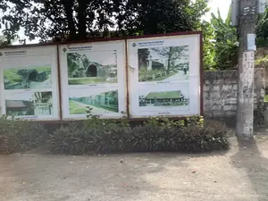 Bản ghi văn bản từ các tấm biển thông tin:。Phía trên cùng: 'TRUNG TÂM BẢO TỒN DI SẢN THÀNH NHÀ HỒ' và 'CONSERVATION CENTER FOR HO CITADEL'.。Chú thích cho các hình ảnh (từ trái qua phải, từ trên xuống dưới): 'CỔNG TÂY', 'NỀN CỔ PHỦ QUỐC TƯỚNG', 'CỔNG ĐÔNG', 'TƯỜNG THÀNH ĐÔNG', 'TIỀU SANH CHƯƠNG', 'ĐÌNH ĐÔNG MÔN'.。Trên cột điện có các ký tự '1.3' và 'T8'.。Mô tả cảnh quan và sự vật:。Bức ảnh chụp một tấm biển thông tin lớn đặt ngoài trời, có thể là ở ven đường gần khu di tích Thành nhà Hồ. Tấm biển này bao gồm nhiều bảng pa-nô, trên đó có dán các bức ảnh giới thiệu về các địa điểm khác nhau trong khu di tích như cổng thành bằng đá, tường thành, nền móng của một công trình kiến trúc cổ và một ngôi đình.。Phía trước tấm biển có một hàng cây bụi trang trí. Bên phải là một cột điện bê tông và một bức tường thấp xây bằng gạch. Phía sau là cây cối xanh tốt, có thể nhận ra một cây lớn và bụi chuối. Mặt đất là nền đất, không lát gạch. Không có người hay hoạt động nào đang diễn ra trong ảnh.。（日本語訳）。案内板からのテキスト記録：。最上部：「TRUNG TÂM BẢO TỒN DI SẢN THÀNH NHÀ HỒ」および「CONSERVATION CENTER FOR HO CITADEL」。写真のキャプション（左から右へ、上から下へ）：「西門」、「国相府古跡」、「東門」、「東城壁」、「蕭笙亭」、「東門亭」。電柱には「1.3」と「T8」の文字がある。風景と物体の説明：。写真は、屋外に設置された大きな案内板を撮影したもので、おそらく胡朝の城塞遺跡近くの道端にある。この案内板は複数のパネルで構成されており、そこには石造りの城門、城壁、古い建築物の基礎、そして一つの亭（てい）など、遺跡内の様々な場所を紹介する写真が貼られている。案内板の前には装飾用の低木が並んでいる。右側にはコンクリートの電柱とレンガで造られた低い壁がある。後ろには青々とした木々があり、大きな木とバナナの茂みが見て取れる。地面は土であり、タイル張りではない。写真には人や活動の様子は写っていない。