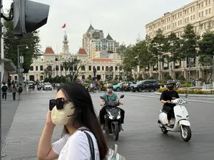 Một cảnh đường phố tại một thành phố lớn ở Việt Nam. Ở phía trước là một phụ nữ trẻ tóc đen, đeo kính râm và khẩu trang. Trên đường phố có nhiều xe máy và ô tô đang di chuyển. Nổi bật ở phía sau là tòa nhà Ủy ban Nhân dân Thành phố Hồ Chí Minh với kiến trúc kiểu Pháp và lá cờ Việt Nam đang bay trên nóc. Nhiều người đi bộ và các tòa nhà hiện đại khác cũng xuất hiện trong khung cảnh.。（日本語訳）。ベトナムのある大都市の街頭風景。手前には、黒髪でサングラスとマスクを着用した若い女性がいます。路上には多くのバイクや車が走っています。後方に目立つのは、フランス様式の建築であるホーチミン市人民委員会庁舎で、屋上にはベトナム国旗がはためいています。多くの歩行者や他の近代的な建物も風景の中に映っています。