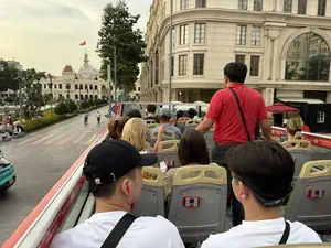 Bức ảnh chụp từ tầng trên của một chiếc xe buýt du lịch hai tầng đang di chuyển trên đường phố ở Thành phố Hồ Chí Minh. Nhiều du khách đang ngồi trên ghế để tham quan, một số người đang đeo tai nghe màu đỏ để nghe thuyết minh. Ở phía xa là tòa nhà Trụ sở Ủy ban Nhân dân Thành phố Hồ Chí Minh với lá cờ Việt Nam đang bay trên đỉnh. Một người đàn ông mặc áo đỏ, có thể là hướng dẫn viên du lịch, đang đứng trên xe. Khung cảnh xung quanh là những tòa nhà mang kiến trúc kiểu Pháp, cây xanh và người đi bộ, đi xe máy trên đường.。（日本語訳）。ホーチミン市の路上を走行中の2階建て観光バスの上階から撮影された写真です。多くの観光客が観光のために座席に座っており、何人かは解説を聞くために赤いヘッドフォンをしています。遠方にはホーチミン市人民委員会庁舎の建物があり、その頂上にはベトナム国旗がはためいています。赤いシャツを着た男性、おそらくツアーガイドが、バスの上に立っています。周囲の風景は、フランス風建築の建物、緑の木々、そして道路上の歩行者やバイクです。