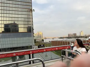 Bức ảnh được chụp từ tầng trên của một chiếc xe buýt du lịch hai tầng, nhìn ra quang cảnh thành phố ven sông. Một người phụ nữ châu Á với mái tóc dài màu đen, mặc áo phông trắng, đang ngồi và chăm chú nhìn về phía trước. Cô ấy đang giữ một chiếc điện thoại di động được gắn trên một thiết bị. Bên trái là một tòa nhà cao tầng hiện đại với mặt tiền hoàn toàn bằng kính. Phía xa là một con sông rộng và đường chân trời của thành phố với nhiều tòa nhà cao tầng dưới bầu trời có mây. Dưới mặt đất, dọc theo bờ sông là một công viên xanh với nhiều người đang đi dạo và thư giãn.。（日本語訳）。長い黒髪で白いTシャツを着たアジア人女性が座って、前方をじっと見つめている。彼女は、ある装置に取り付けられた携帯電話を持っている。左側には、全面ガラス張りの近代的な高層ビルがある。地上では、川岸に沿って緑豊かな公園があり、多くの人々が散歩したりリラックスしたりしている。