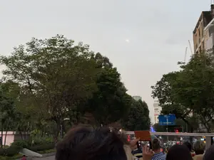 Bức ảnh chụp khung cảnh thành phố vào lúc chạng vạng, nhìn từ một vị trí trên cao, có thể là trên một chiếc xe buýt du lịch. Bầu trời có màu xám nhạt với một vầng trăng mờ. Phía trước là nhiều người, trong đó có một người đang dùng điện thoại thông minh với vỏ màu nâu để chụp ảnh. Hai bên là những hàng cây xanh tươi tốt. Ở phía xa, có thể thấy các tòa nhà cao tầng, cần cẩu xây dựng và một biển báo giao thông màu xanh không rõ nội dung.。（日本語訳）。黄昏時の街の風景を撮影した写真で、高い位置、おそらく観光バスの上から見たものである。前方には多くの人がおり、その中には茶色いケースのスマートフォンを使って写真を撮っている人がいる。両側には青々とした並木がある。