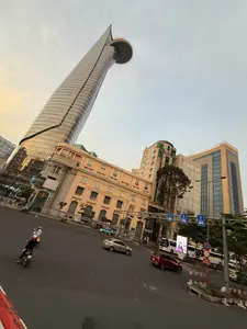 Bức ảnh chụp một góc phố tại Thành phố Hồ Chí Minh, nổi bật với tòa nhà chọc trời Bitexco Financial Tower có hình dáng độc đáo như một búp sen và có một sân bay trực thăng ở gần đỉnh. Xung quanh là các tòa nhà khác với kiến trúc đa dạng, từ một tòa nhà cổ màu vàng mang phong cách Pháp đến các cao ốc văn phòng hiện đại. Dưới đường, dòng xe cộ gồm xe máy, ô tô và xe buýt đang di chuyển tấp nập tại một ngã tư. Bầu trời có mây, cho thấy thời điểm là lúc hoàng hôn hoặc bình minh, tạo nên một khung cảnh đô thị vừa hiện đại vừa năng động.。（日本語訳）。ホーチミン市のある街角を撮影した写真で、蓮の蕾のようなユニークな形をし、頂上近くにヘリポートがある超高層ビル「ビテクスコ・フィナンシャルタワー」が際立っている。周囲には、フランス風の黄色い古い建物から近代的なオフィスビルまで、様々な建築様式の他の建物がある。道路では、バイク、自動車、バスなどの交通が交差点でひしめき合っている。空には雲があり、夕暮れ時か夜明け時であることを示しており、現代的かつダイナミックな都市の風景を作り出している。