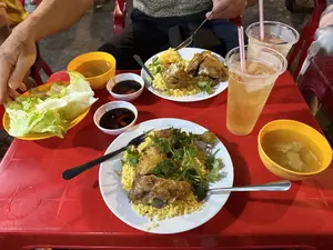 Trên một chiếc bàn nhựa màu đỏ, có hai đĩa cơm gà. Cơm có màu vàng, phía trên là những miếng gà rán và rau mùi. Bên cạnh mỗi đĩa cơm là một bát canh nhỏ màu vàng và một chén nước chấm. Ngoài ra còn có một đĩa rau xà lách và hai ly trà đá bằng nhựa trong suốt với ống hút màu hồng. Hai người đang ngồi ăn tại bàn.。（日本語訳）。赤いプラスチックのテーブルの上に、コムガー（チキンライス）が2皿あります。ご飯は黄色で、その上にはフライドチキンの切り身とパクチーが乗っています。それぞれの皿の横には、黄色い小さなスープ椀とタレの小鉢が一つずつあります。その他に、レタスの皿が1枚と、ピンクのストローが差さった透明なプラスチック製のグラスに入ったアイスティーが2杯あります。二人の人がテーブルについて食事をしています。