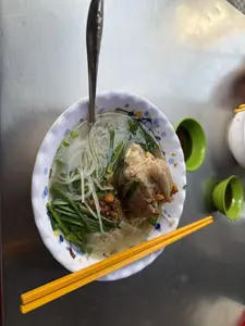 Một tô bún nước lèo được đặt trên một chiếc bàn inox. Trong tô có bún sợi trắng, một cục giò heo lớn, rất nhiều hành hẹ cắt khúc và một ít hành phi. Một chiếc thìa inox đang cắm trong tô, và một đôi đũa màu vàng được đặt ngang trên miệng tô. Bên cạnh tô có hai chiếc chén nhỏ màu xanh lá cây.。（日本語訳）。一杯のブンヌックレオがステンレス製のテーブルの上に置かれています。丼の中には、白いブン、大きな豚足の塊一つ、たくさんのぶつ切りにしたネギとニラ、そして少しのフライドオニオンが入っています。一本のステンレス製のスプーンが丼に刺さっており、一膳の黄色い箸が丼の縁に横向きに置かれています。丼の隣には、緑色の小さな小鉢が二つあります。