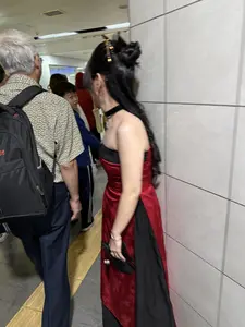 Một người phụ nữ có mái tóc đen dài được búi cao và cài trâm, mặc một chiếc váy dài cách tân màu đỏ và đen, thiết kế hở vai. Cô đeo vòng cổ choker màu đen và tay cầm một chiếc quạt màu đen. Bên cạnh cô là một người đàn ông lớn tuổi tóc bạc, mặc áo sơ mi hoạ tiết và đeo ba lô. Xung quanh có những người khác trong một hành lang có tường ốp gạch.。（日本語訳）。長い黒髪を高く結い上げ、かんざしを挿した女性が、赤と黒のオフショルダーデザインのモダンなロングドレスを着ている。彼女は黒いチョーカーをつけ、黒い扇子を手に持っている。彼女の隣には、白髪の年配の男性がおり、柄物のシャツを着てバックパックを背負っている。周りには、タイル張りの壁の廊下に他の人々がいる。