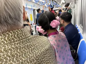 Bức ảnh chụp từ phía sau bên trong một toa tàu điện ngầm đông đúc. Ở phía trước, một người lớn tuổi với mái tóc bạc đang ngồi, mặc một chiếc áo sơ mi có hoa văn hình học. Bên cạnh họ là một người phụ nữ với mái tóc đen được búi lên và cài nhiều bông hoa màu hồng. Cô ấy mặc một chiếc áo dài màu hồng có hoa văn. Xung quanh có nhiều hành khách khác, một số đang đứng và vịn vào tay nắm màu xanh trên tàu.。（日本語訳）。混雑した地下鉄の車両内を後ろから撮影した写真です。前方には、白髪の年配の方が座っており、幾何学模様のシャツを着ています。その隣には、黒髪をアップにし、ピンク色の花をたくさん挿した女性がいます。彼女は模様の入ったピンク色のアオザイを着ています。周りには他の乗客も多く、何人かは立って車内の青いつり革に掴まっています。