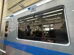 Bức ảnh chụp một toa tàu điện màu bạc có sọc xanh dương đang dừng tại một nhà ga. Nhìn qua cửa sổ lớn, có thể thấy cảnh sinh hoạt bên trong toa tàu với nhiều hành khách. Một số người đang đứng, tay bám vào các vòng tay vịn treo trên cao, trong khi những người khác ngồi trên ghế. Trong số các hành khách, có thể nhận ra một người đội mũ và một người phụ nữ đang đứng gần cửa sổ nhỏ hơn. Ánh đèn điện bên trong toa tàu và hình ảnh phản chiếu của kết cấu mái nhà ga trên bề mặt kính tạo nên một khung cảnh phức hợp, thể hiện một khoảnh khắc thường ngày trên phương tiện giao thông công cộng.。（日本語訳）。青い帯の入った銀色の電車が駅に停車している写真です。大きな窓からは、多くの乗客がいる車内の様子が見えます。何人かは立ち、高い位置に吊り下げられた吊り革に掴まっており、他の人々は座席に座っています。乗客の中には、帽子をかぶった人と、より小さい窓の近くに立っている女性が見て取れます。車内の電灯の光と、ガラス面に反射した駅の屋根の構造が複雑な光景を作り出し、公共交通機関における日常の一コマを表しています。