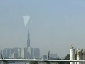 Bức ảnh chụp một khung cảnh thành phố hiện đại, mờ ảo trong sương mù. Ở phía xa, một tòa nhà chọc trời cao vút, có khả năng là Landmark 81, nổi bật giữa các tòa nhà cao tầng khác. Phía trước là một con sông và các công trình công nghiệp như cần cẩu và silo. Toàn bộ khung cảnh bị bao phủ bởi một lớp sương dày, khiến các chi tiết trở nên không rõ ràng.。（日本語訳）。写真は、霧でぼやけた現代的な都市の風景を写している。遠方には、ランドマーク81と思われるそびえ立つ超高層ビルが、他の高層ビルの中で際立っている。手前には川と、クレーンやサイロのような産業施設がある。風景全体が厚い霧の層に覆われ、細部が不鮮明になっている。